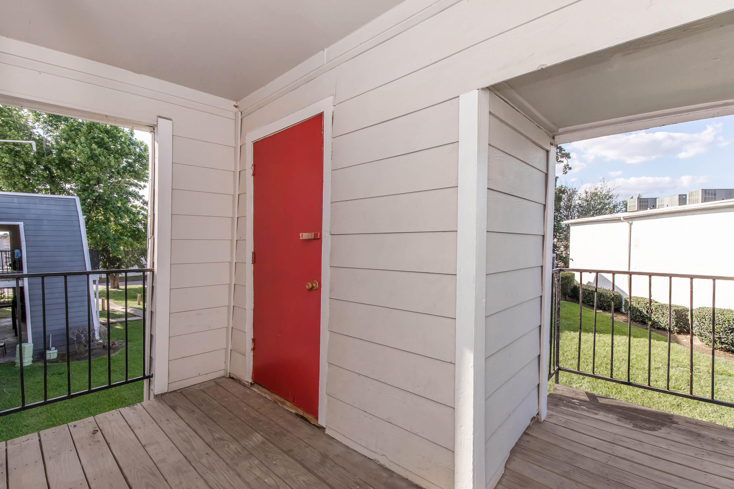A view of a covered balcony with wooden flooring. On one side, there is a bright red door, while the other side opens to an outdoor area with green grass and trees in the background. The walls are white, creating a clean and simple aesthetic.