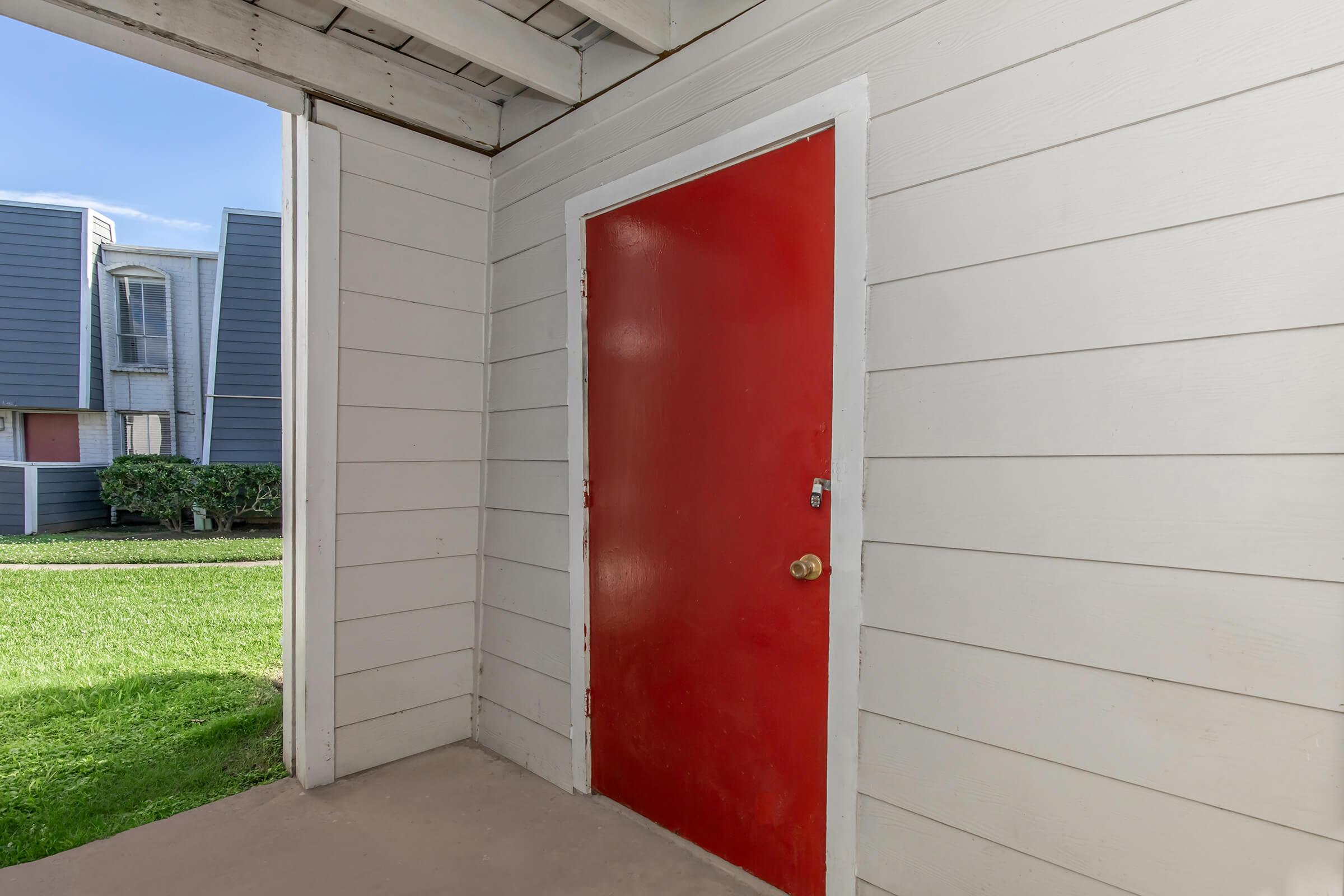 A bright red door stands in a white-paneled exterior, framed by a covered porch. The surrounding area features green grass and glimpses of buildings with varying colors in the background, indicating a residential setting. The lighting suggests a clear, sunny day.