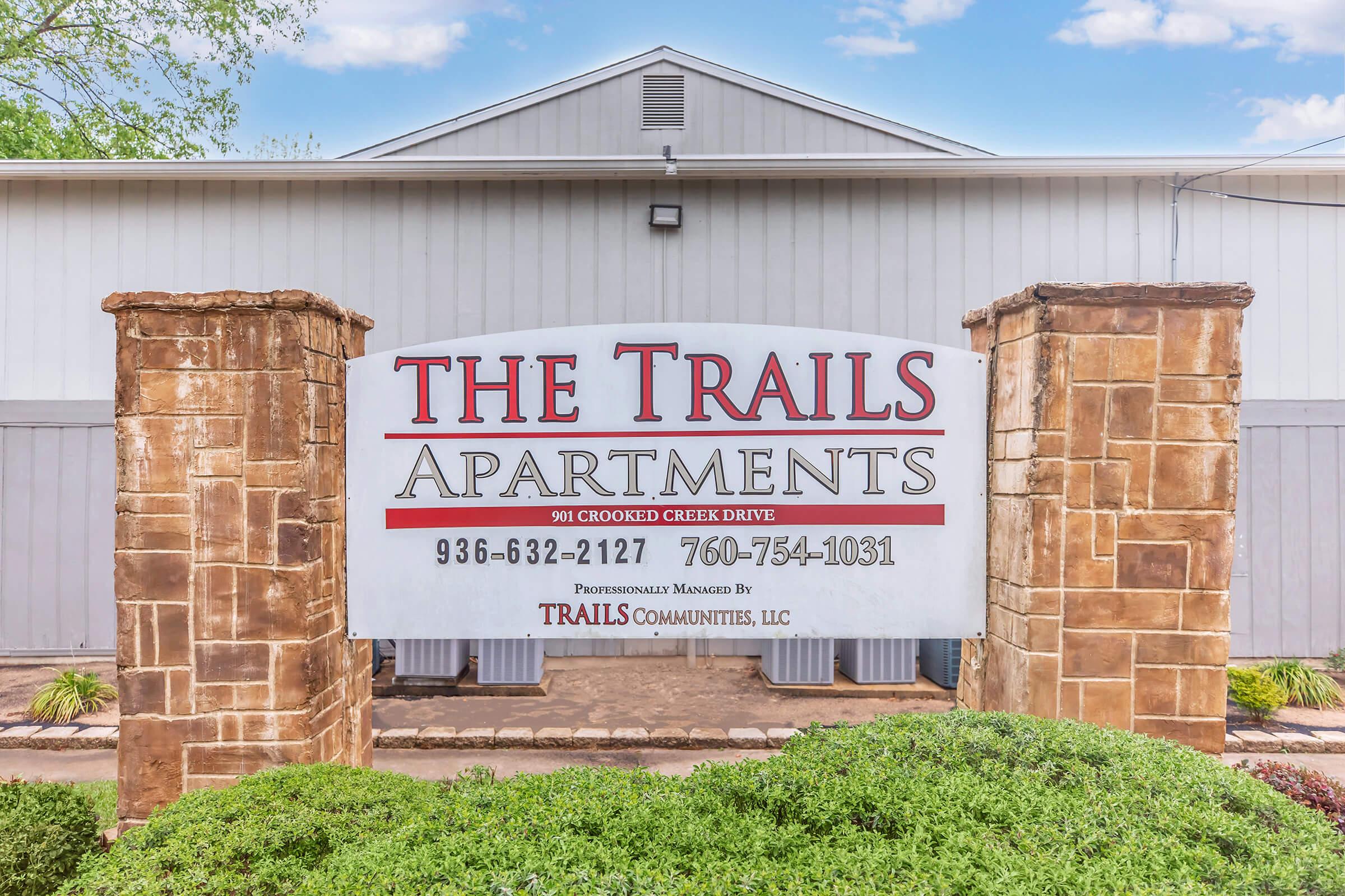 Sign at the entrance of The Trails Apartments, featuring the name prominently displayed along with the address "901 Crooked Creek Drive" and contact numbers "936-632-2127" and "760-754-1031." Surrounded by decorative stone pillars and greenery, set against a clear blue sky.