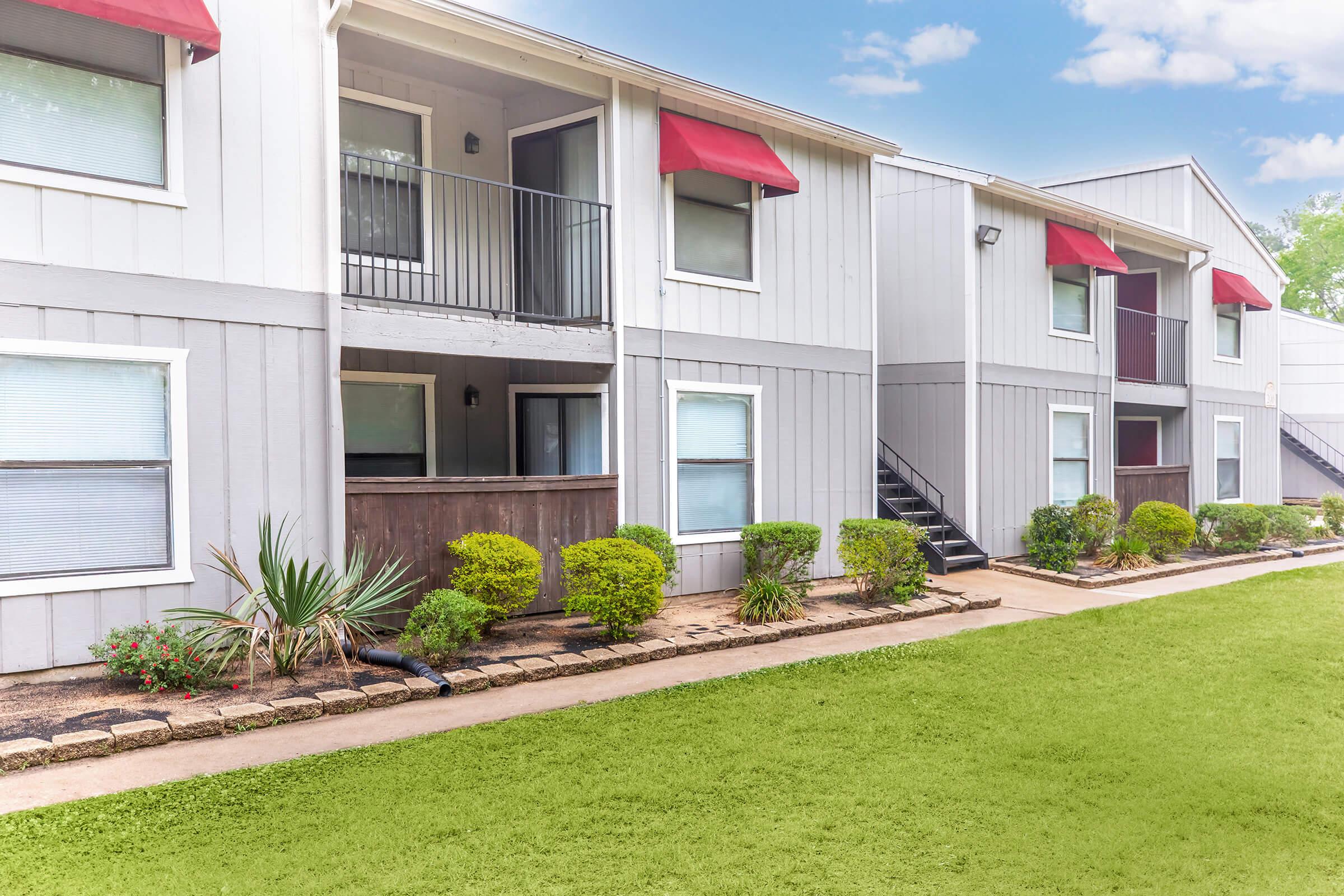 A well-maintained multi-unit apartment complex featuring gray siding, red awnings over windows, and a landscaped path. The lawn is green with shrubs and small plants along the walkway. There are balconies visible on the upper level of the building. Clear skies add to the inviting atmosphere.