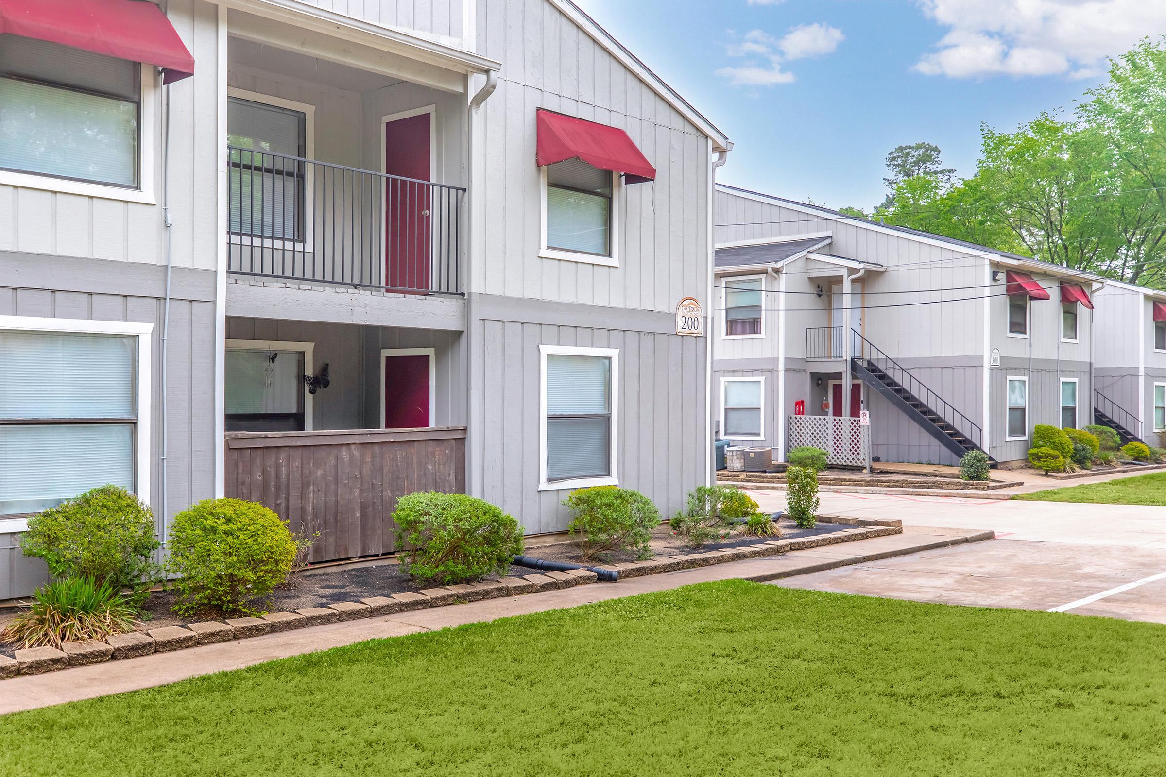 Exterior view of a two-story apartment building with grey siding and red awnings. The ground is paved, and there's a green lawn in front. The building features balconies, windows with sheer curtains, and a staircase leading to the second floor. Trees are visible in the background, indicating a pleasant environment.