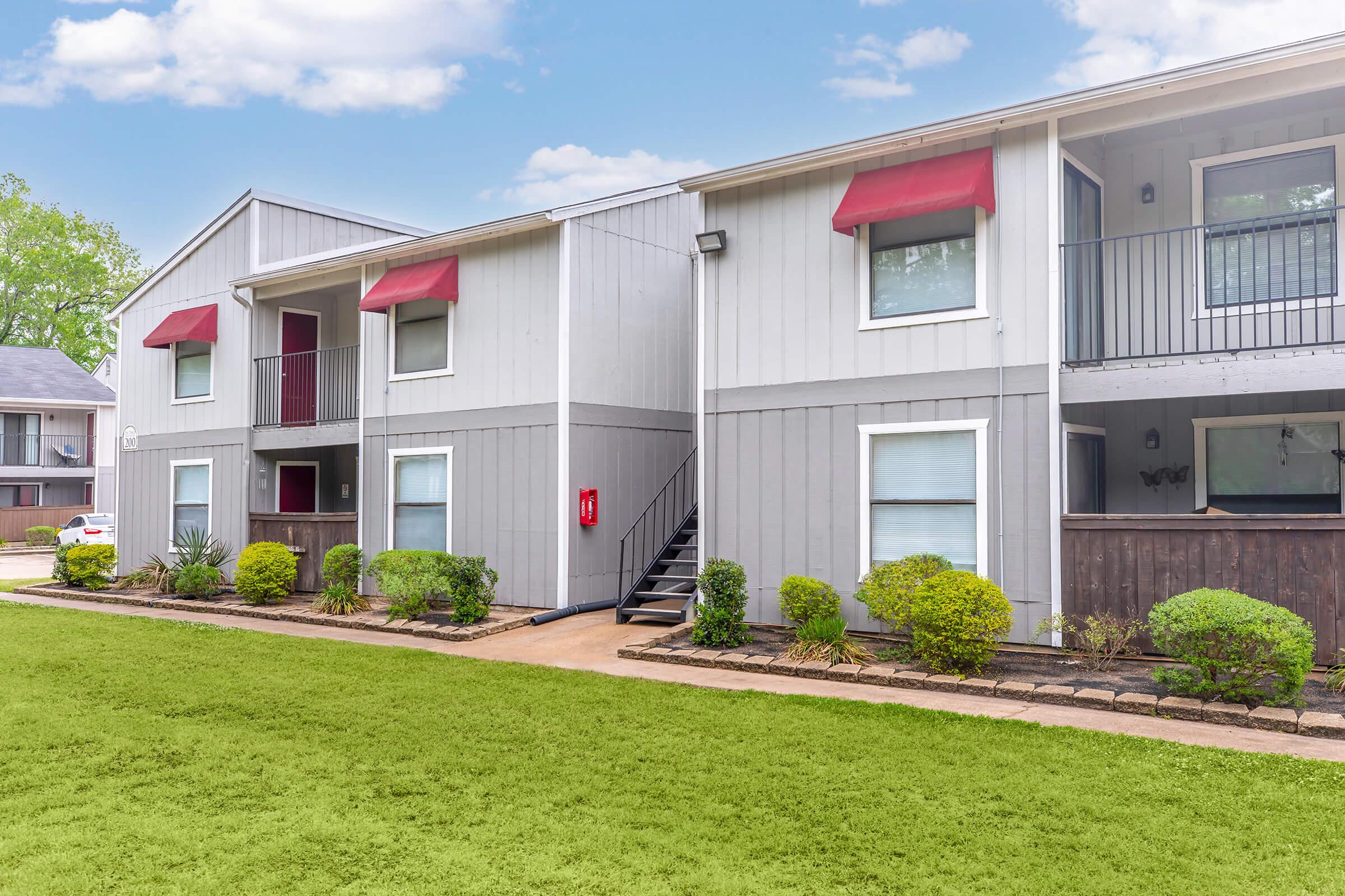 Gray two-story apartment building with red awnings over windows, surrounded by well-maintained shrubs and a green lawn. A set of stairs leads to the upper level, and a fire extinguisher is mounted on the wall. The sky is clear with a few clouds.