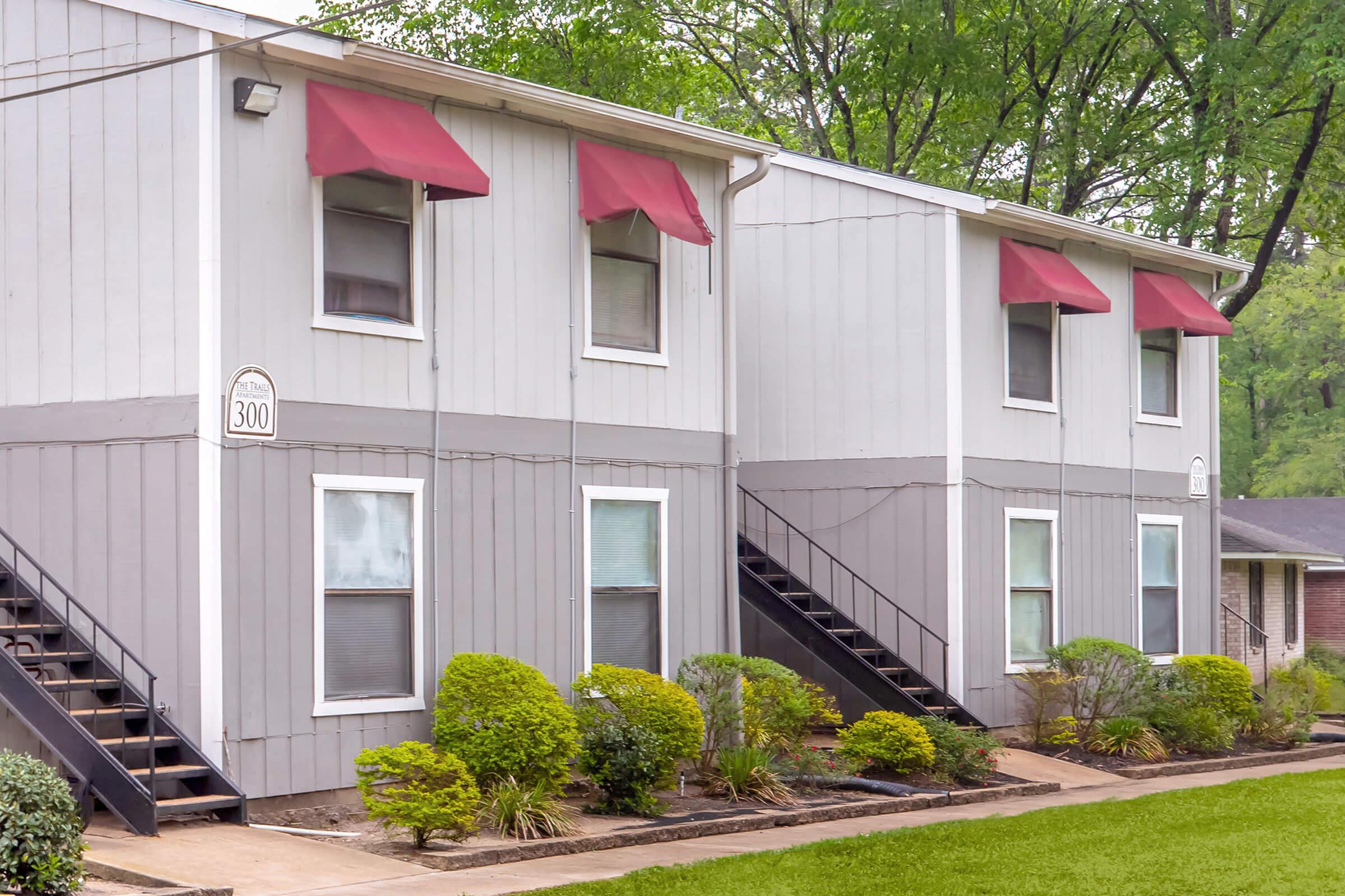 A two-story apartment building featuring a gray exterior, red awnings over the windows, and a small landscaped area with shrubs. There are two sets of stairs leading to the upper floor units, and the building is surrounded by a well-maintained lawn and greenery.