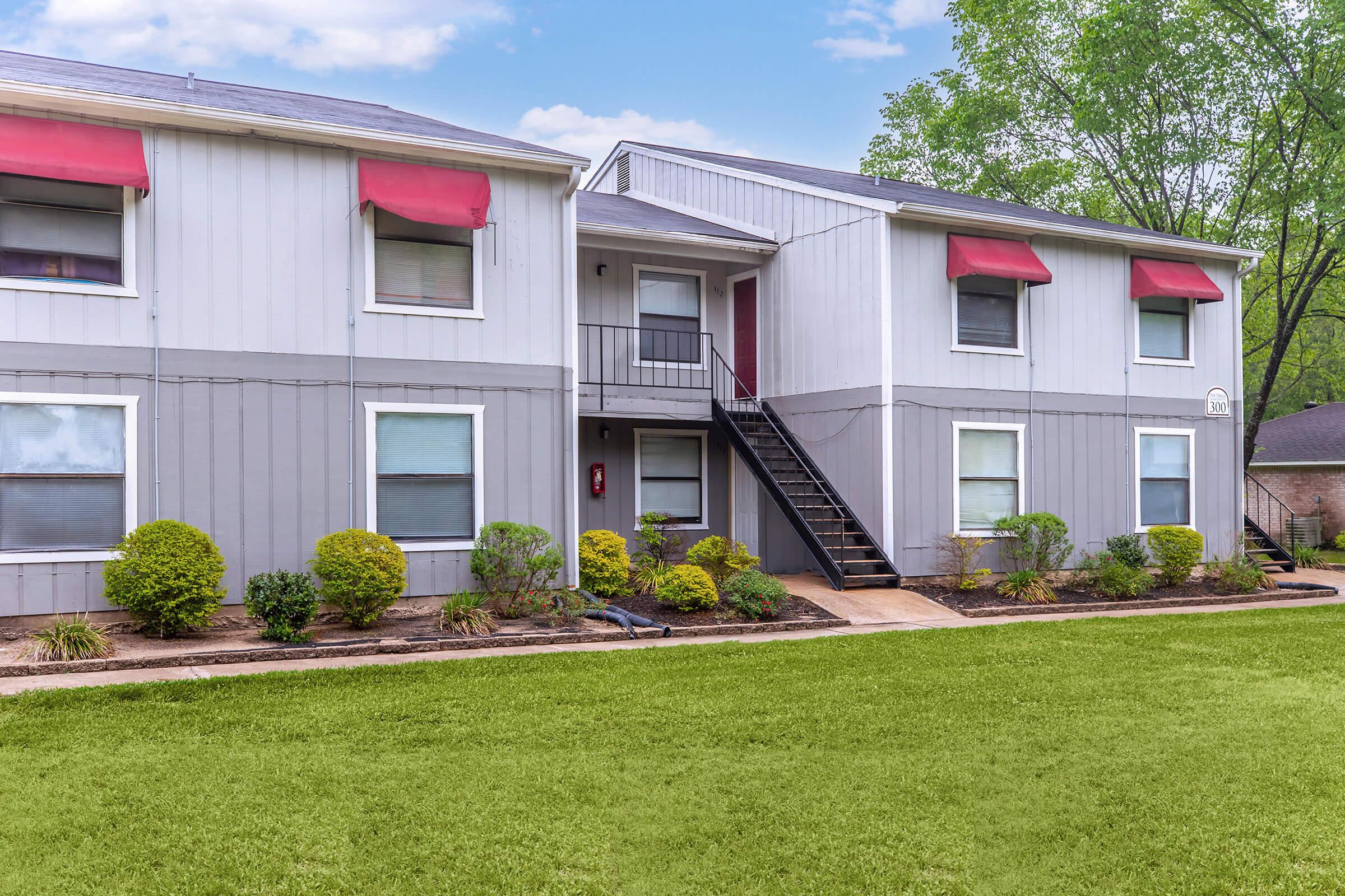 A two-story apartment building with gray siding and red awnings above the windows. The entrance features a staircase leading to the second floor. The front yard is well-maintained with green grass and shrubs, creating a welcoming atmosphere. Trees are visible in the background under a partly cloudy sky.