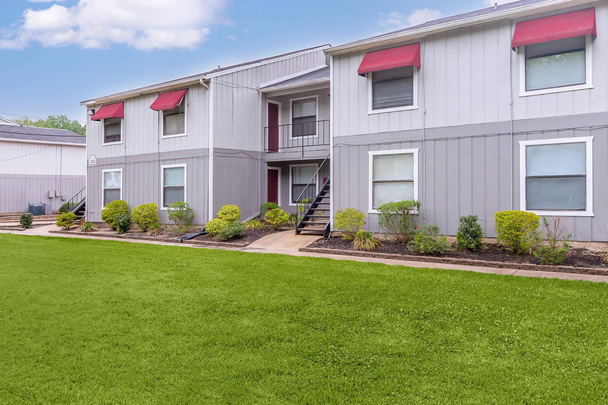 A two-story apartment building with light gray siding, featuring red awnings over the windows. There are manicured shrubs and small plants along a pathway leading to the entrance. The lawn is well-kept with vibrant green grass, and the sky is partly cloudy in the background.