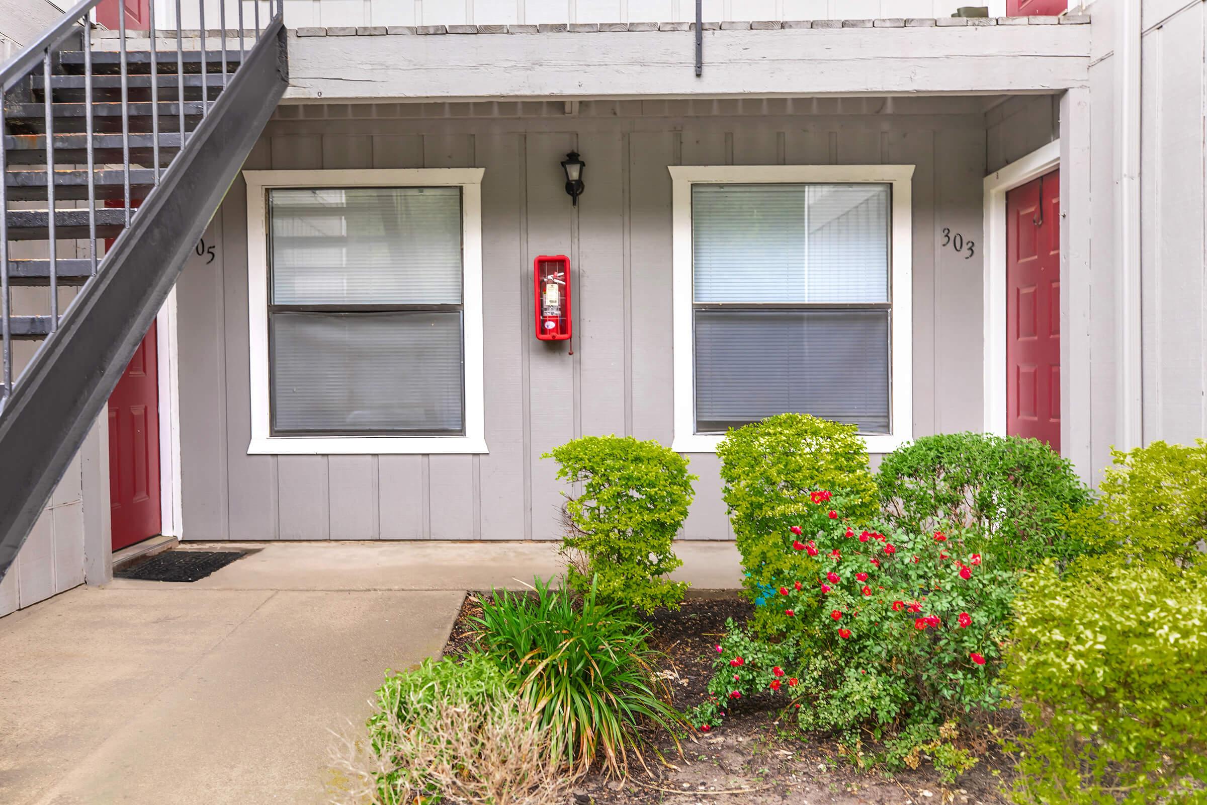 A ground-level view of an apartment entrance showing two windows with blinds, a red fire extinguisher mounted on the wall, and a red door numbered 303. Lush green bushes and colorful flowers are planted in front, with a staircase visible on the left side.