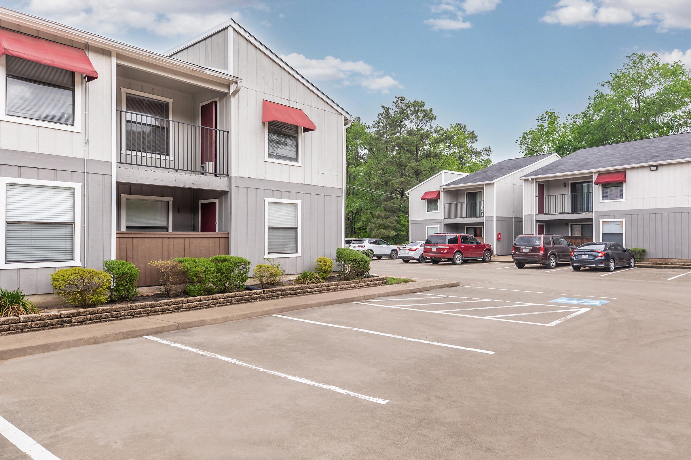 A residential apartment complex featuring two-story buildings with red awnings. The parking lot is visible in the foreground, with marked parking spaces, including a designated spot for disabled parking. Lush greenery surrounds the buildings, and a few cars are parked nearby under a clear blue sky.