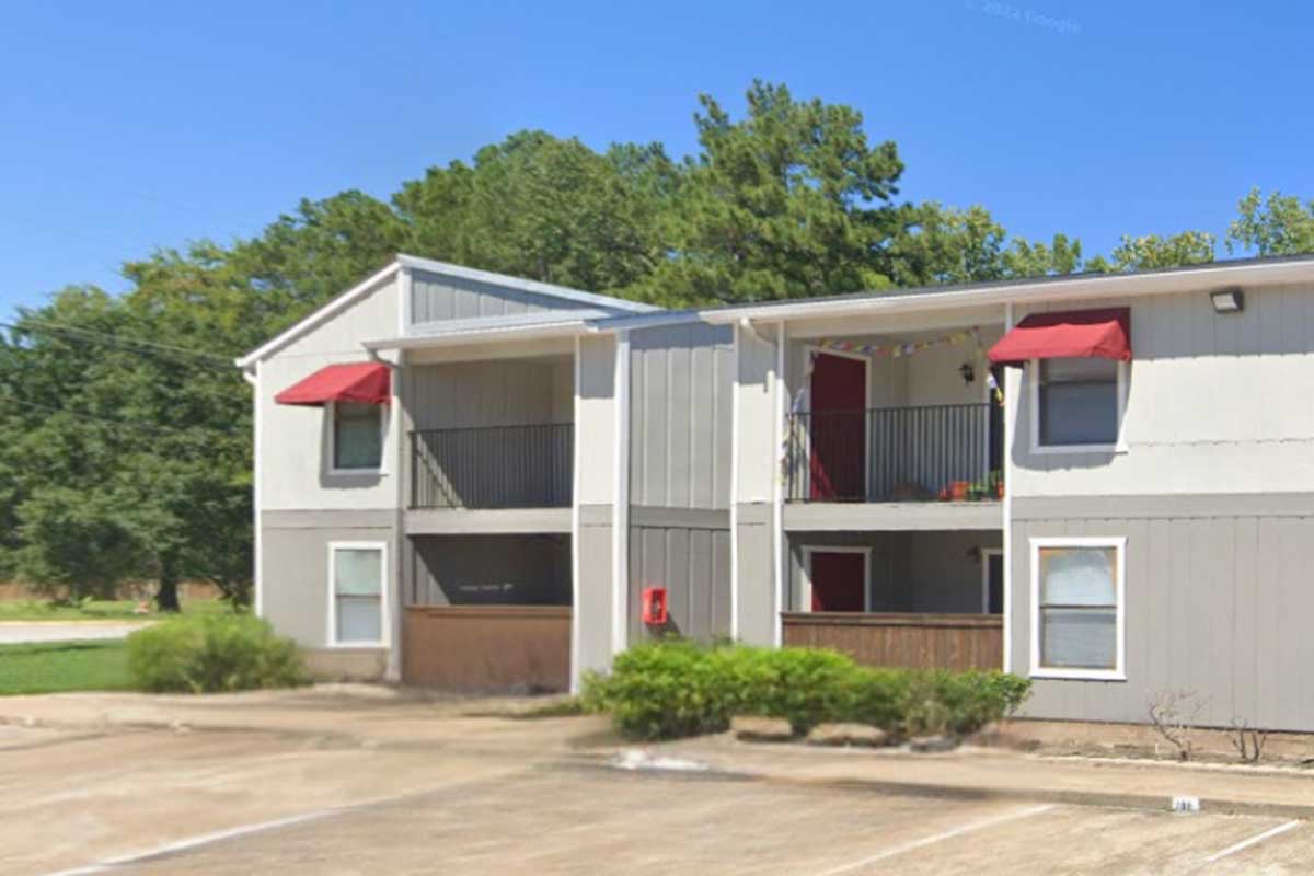 A two-story apartment building with gray siding and red awnings over the balconies. The ground floor features a small patio area, and there's a parking lot in front. Lush green trees are visible in the background under a clear blue sky.