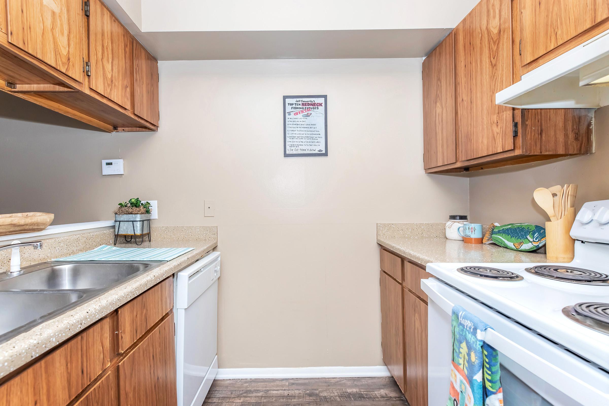 A modern kitchen featuring wooden cabinets, a countertop with a sink, a dishwasher, and a stove. The walls are painted light beige, and a sign is hanging on the wall. A few kitchen utensils and decorative items are placed on the counters, with a colorful towel hanging from the oven.