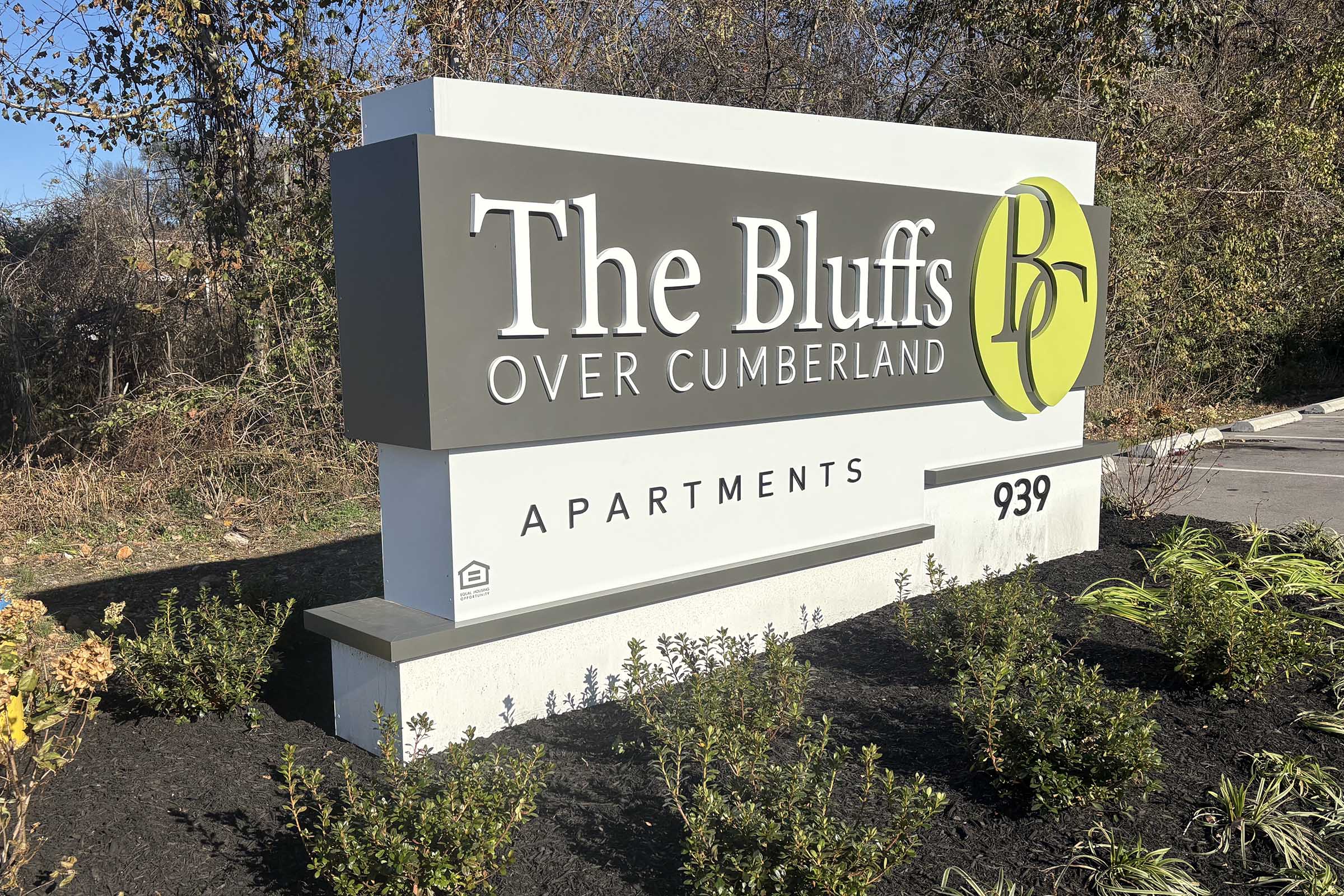 Sign for "The Bluffs Over Cumberland" apartments, displaying the address "939." The sign features a modern design with a green circular logo, set against a natural backdrop of trees and shrubs. The overall appearance suggests a welcoming residential community.