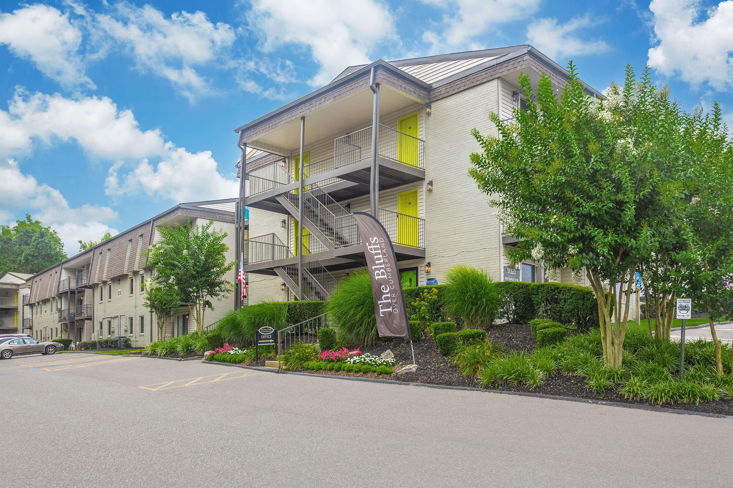 Exterior view of a multi-story apartment building named "The Buffs," featuring a light-colored facade, yellow accents, and balconies. A landscaped area with greenery and flowers is in the foreground, along with parking spaces and a flag/banner displaying the building's name. The sky is blue with scattered clouds.