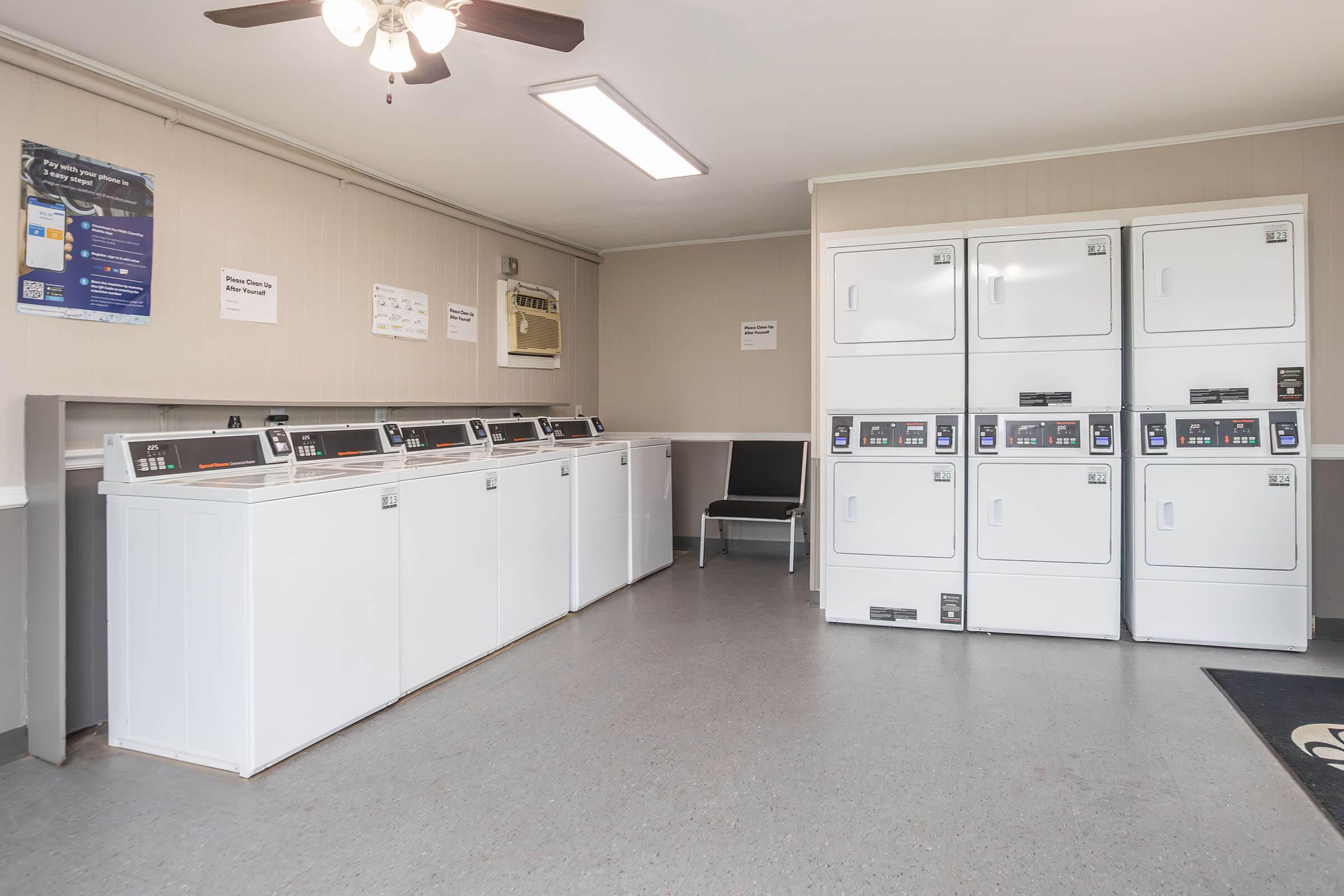 A clean and well-lit laundry room featuring several white washing machines and dryers lined against the wall. There is a ceiling fan above, a wall-mounted air conditioner, and a single chair positioned nearby. Informational signs are posted on the walls. The floor is tiled with a gray finish.