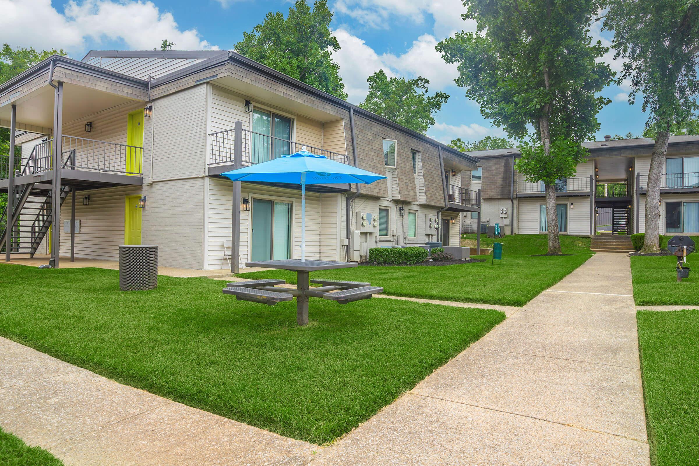 A well-maintained residential complex featuring two buildings with staircases, vibrant green lawns, and a picnic area with a blue umbrella. The scene is bright and sunny, showcasing a peaceful outdoor environment for residents.