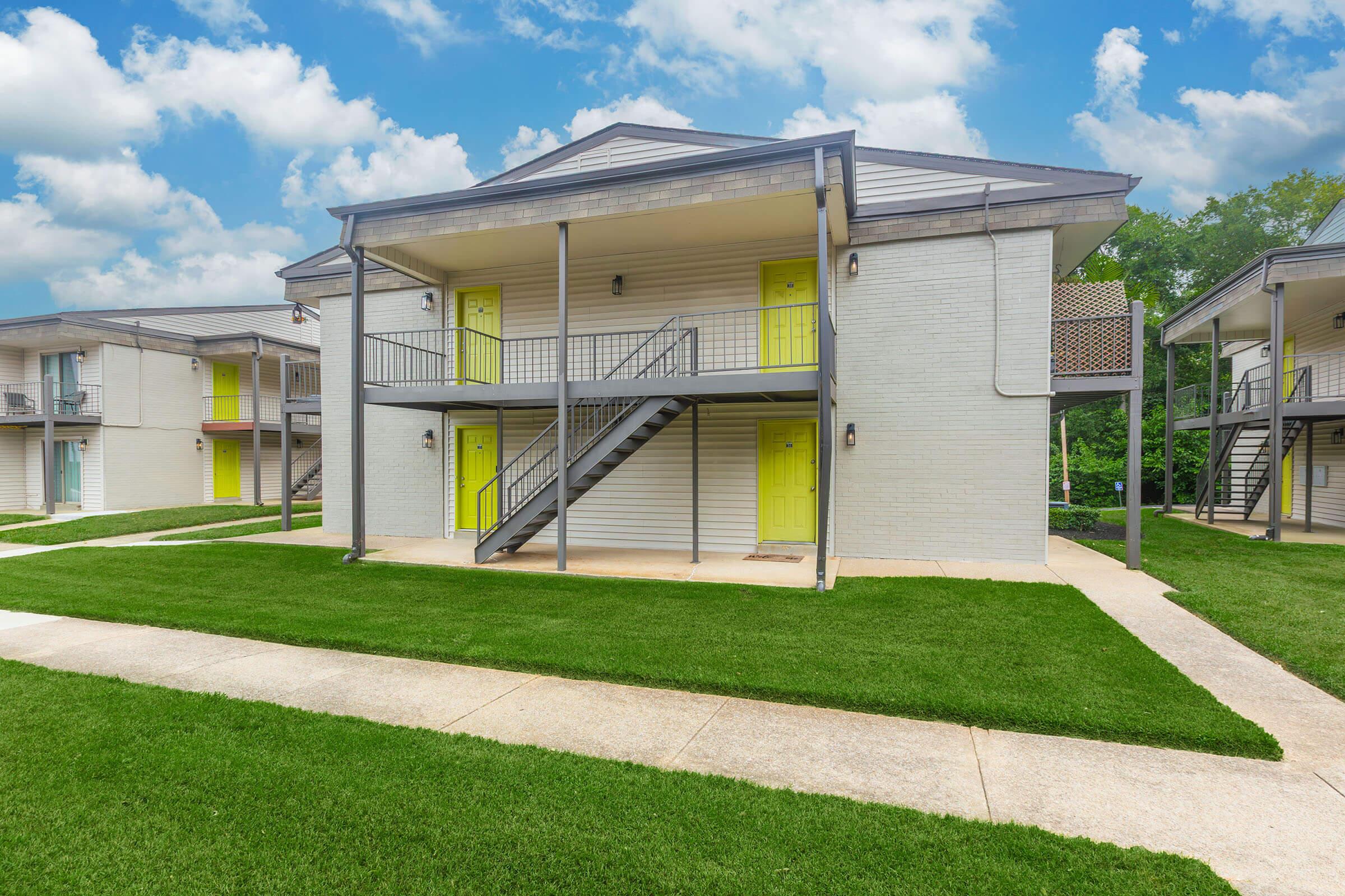A two-story apartment building with a light gray exterior and vibrant yellow doors. The structure features a staircase leading to the second floor. The surrounding area has well-maintained green grass and a concrete walkway, with fluffy clouds visible in a blue sky above.