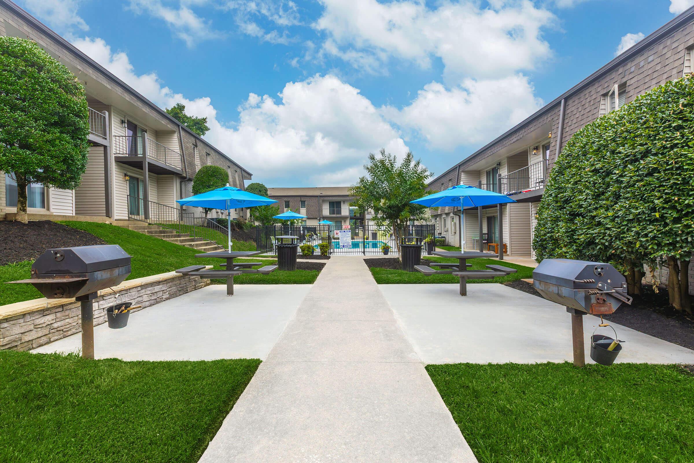 A well-maintained outdoor area featuring a central walkway leading to a swimming pool. Surrounding the walkway are trees and bushes, along with several picnic tables and barbecue grills. Blue umbrellas provide shade over the seating areas, creating an inviting space for relaxation and outdoor activities.