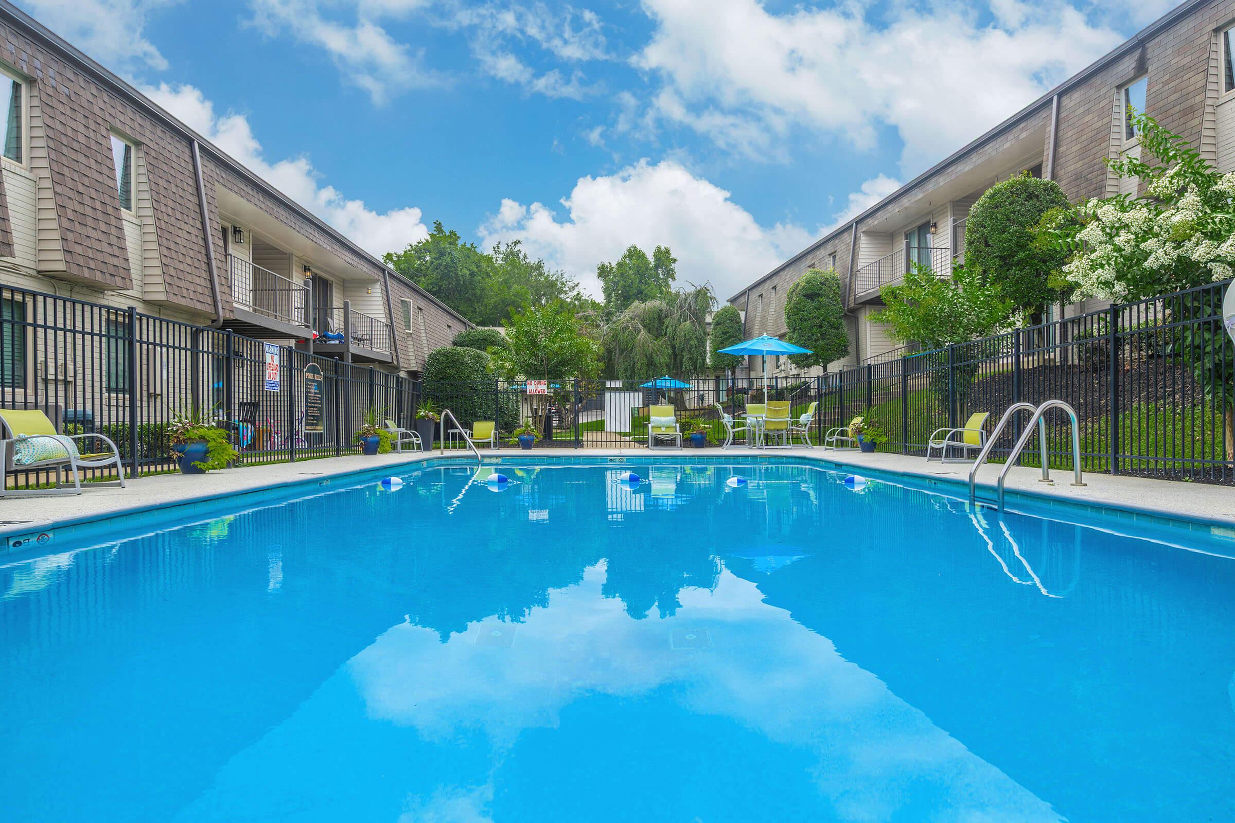 A clear blue swimming pool surrounded by a fenced area, with lounge chairs and umbrellas. Two-story apartment buildings are visible in the background, along with trees and shrubs. The sky is partly cloudy, reflecting on the water's surface.