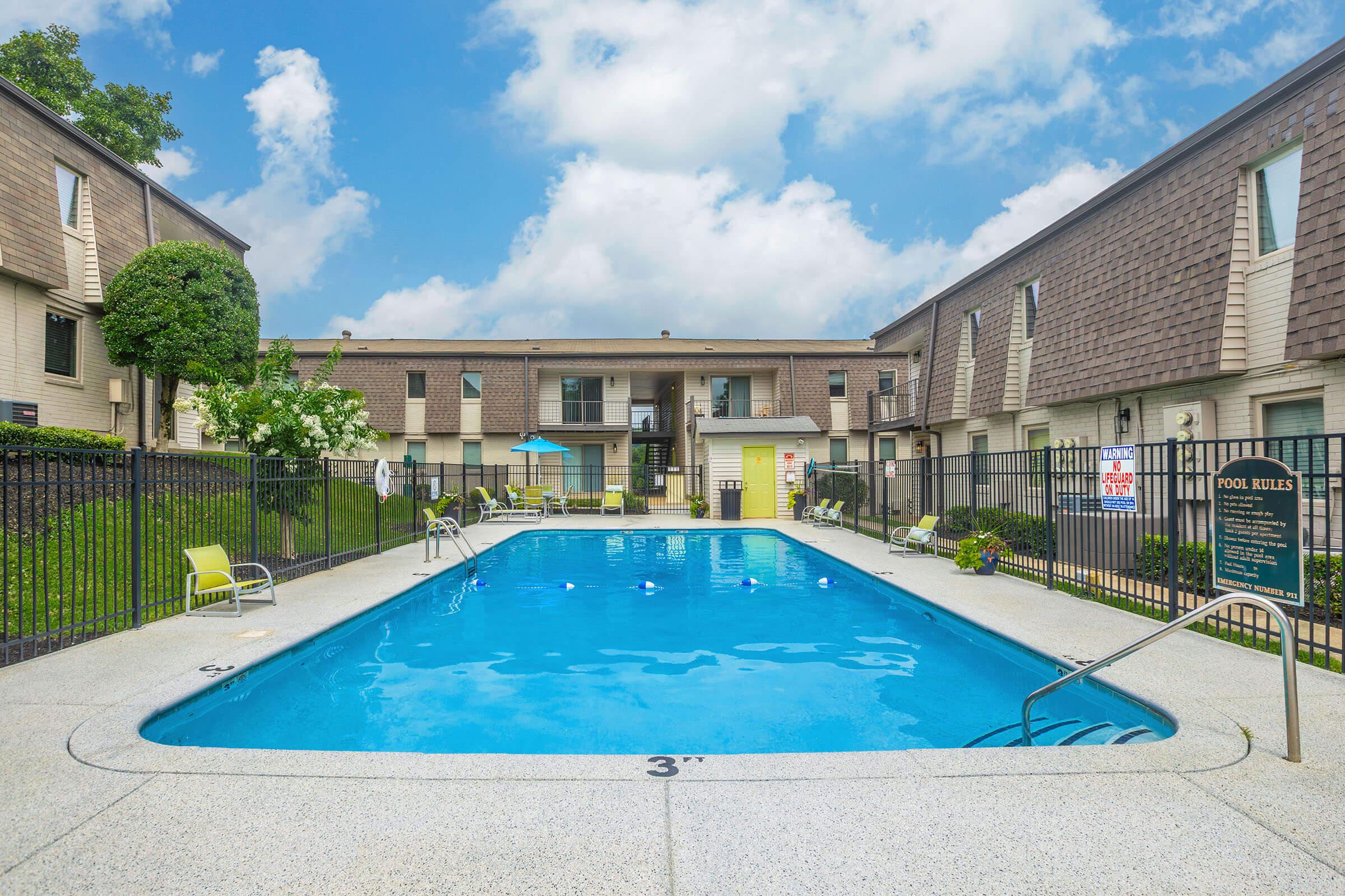 A clear outdoor swimming pool surrounded by a concrete deck, with lounge chairs positioned around it. In the background, two multi-story buildings with balconies are visible, and a green lawn and trees provide some landscaping. The sky is partly cloudy, creating a bright atmosphere.