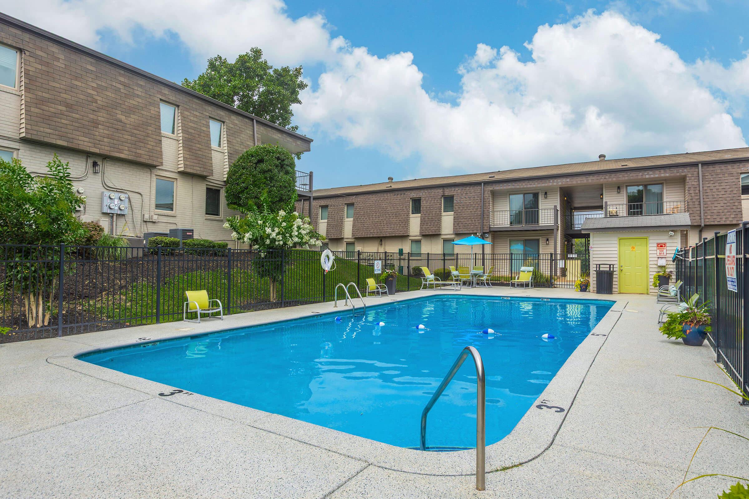 A clear swimming pool surrounded by a deck with lounge chairs, umbrellas, and landscaping. In the background, there are two stories of apartment buildings with a bright yellow door visible. The sky is partly cloudy, creating a pleasant outdoor atmosphere.