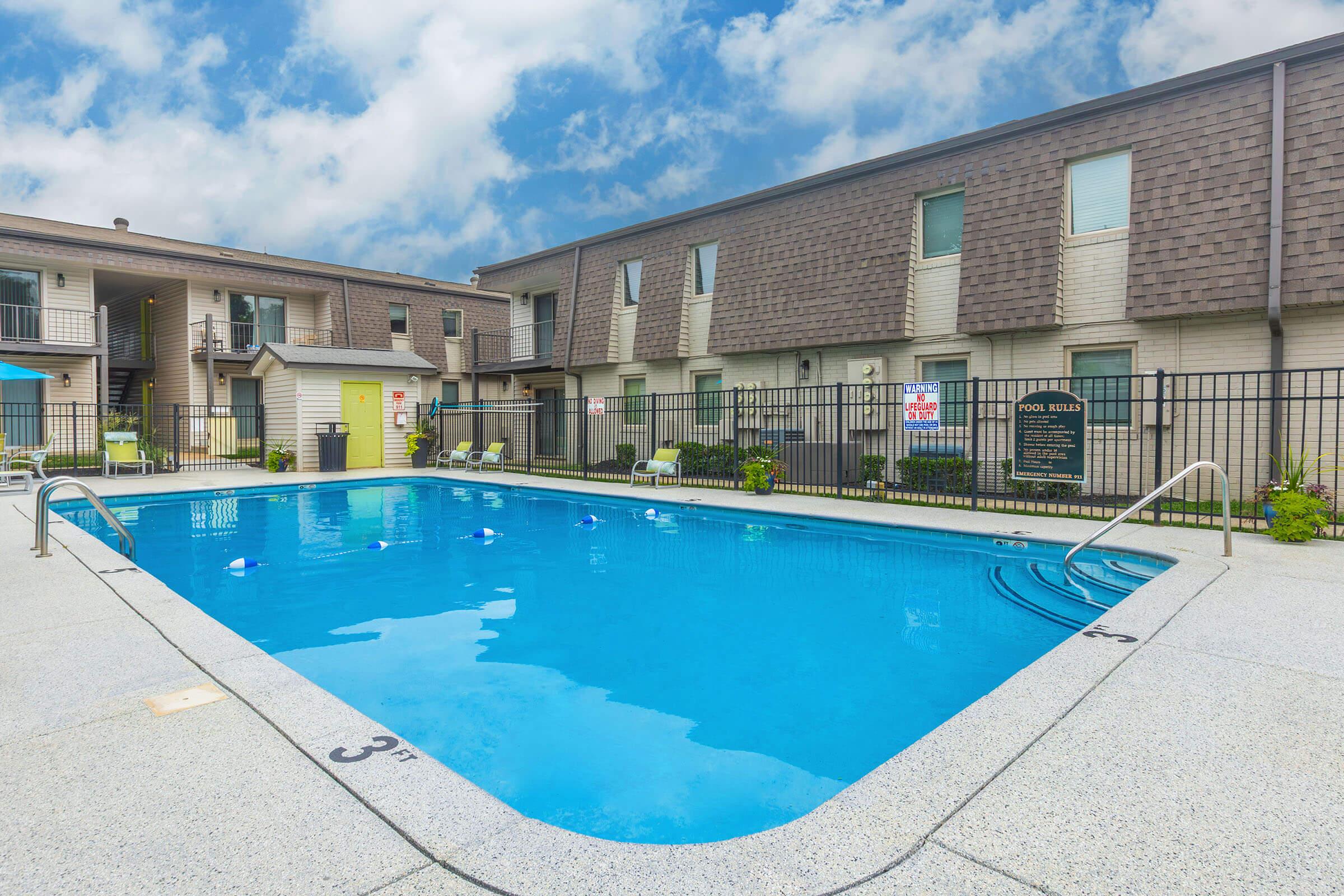 A clear swimming pool in an apartment complex, surrounded by a well-maintained patio area. In the background, two buildings with multiple windows are visible, and a sign indicates the pool rules. Lounge chairs are placed near the pool, and the sky is partly cloudy.