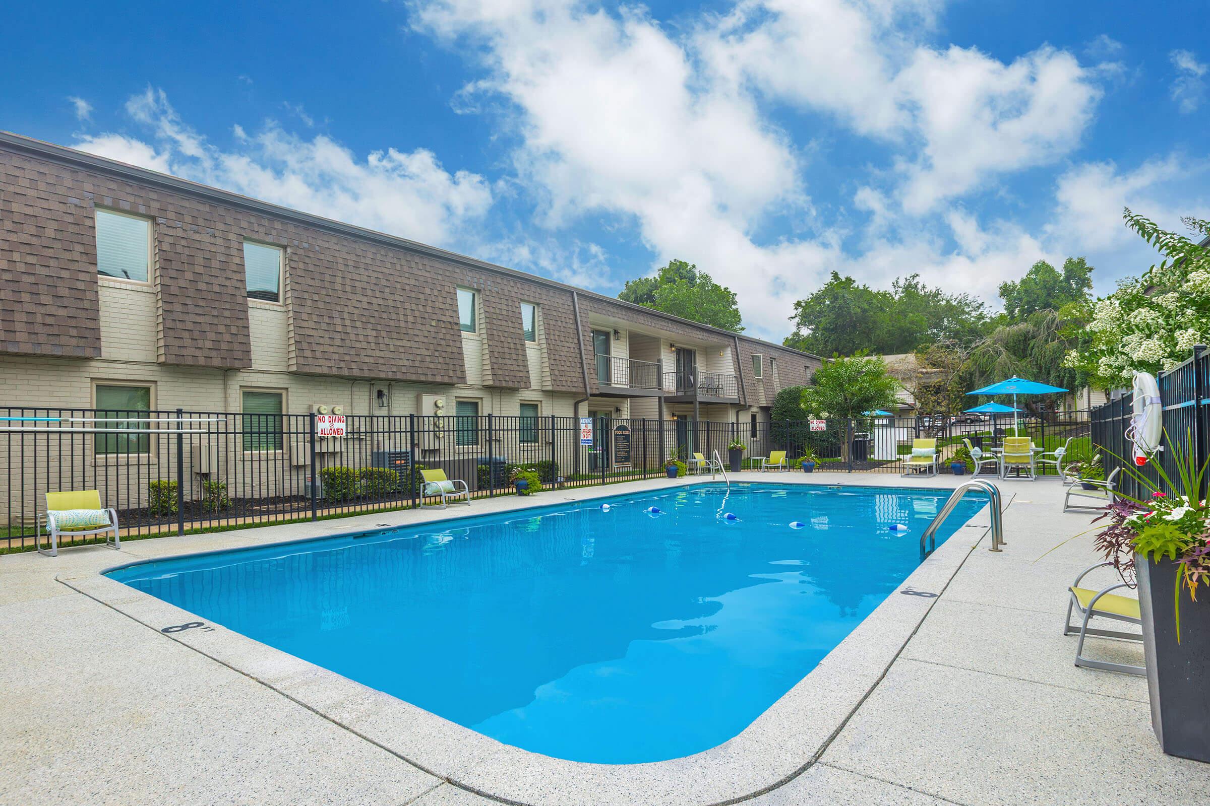 A swimming pool surrounded by lounge chairs and patio tables, located near a multi-story building. Lush greenery and patio umbrellas are visible in the background under a partly cloudy blue sky. The pool area is enclosed with a black fence for security.