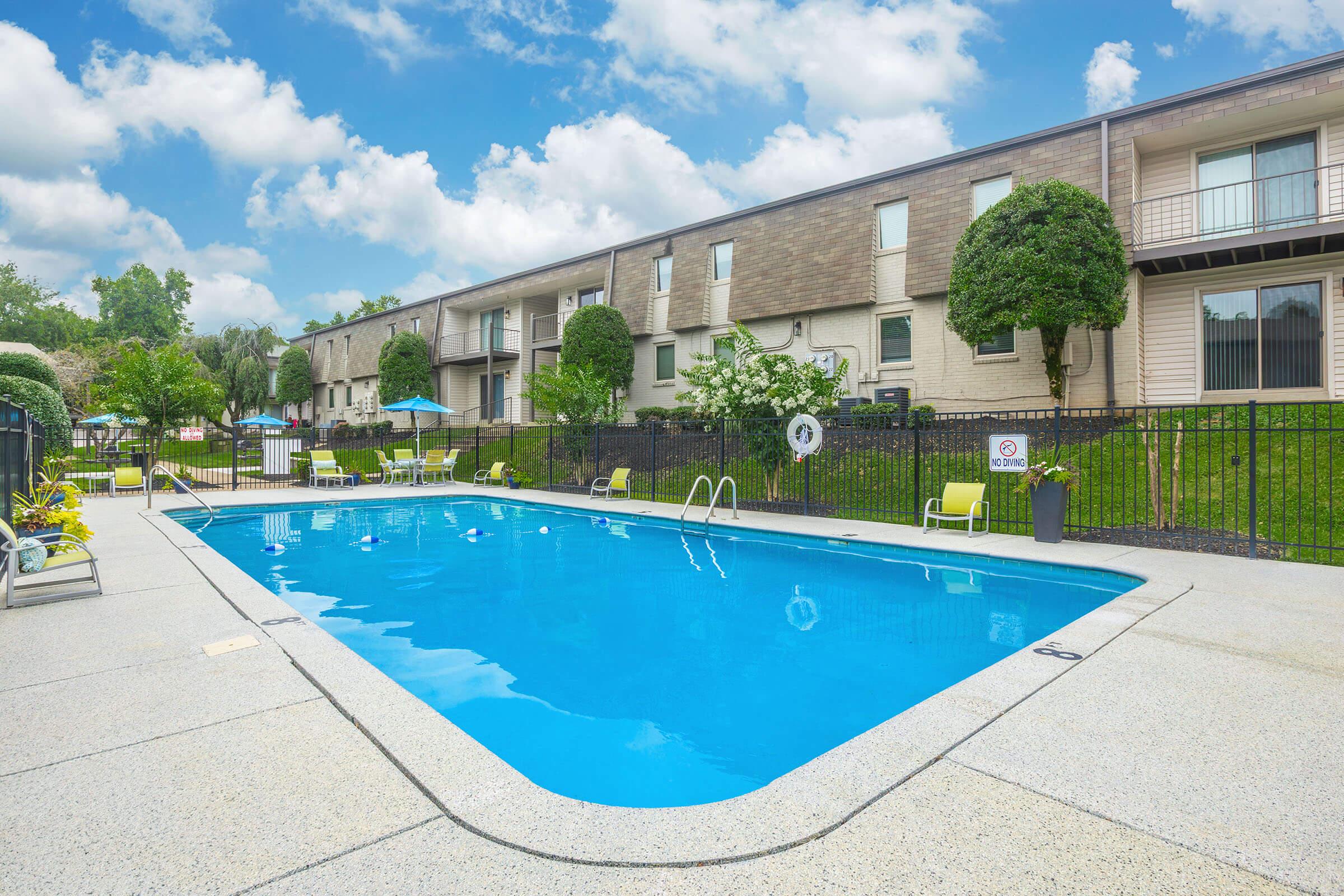 A clear blue swimming pool surrounded by a well-maintained patio area. In the background, there are two-story buildings with balconies and green landscaping. Several lounge chairs are positioned near the pool, and the sky is partly cloudy, creating a bright and inviting atmosphere.