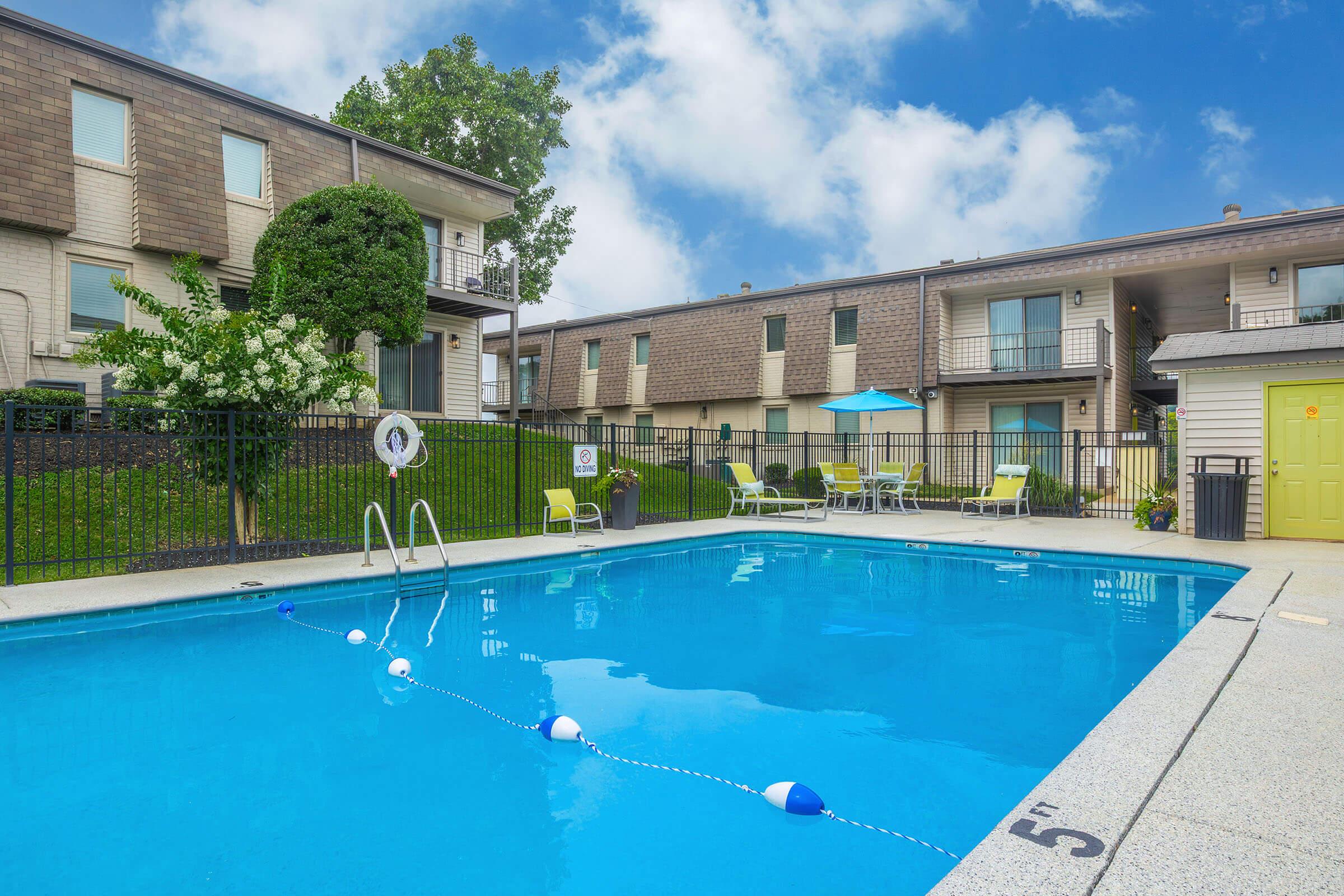 A clear blue swimming pool with a ladder, surrounded by a fenced area. Lounge chairs and an umbrella are placed nearby, with green grass and trees in the background. The sky is partly cloudy, and an apartment building is visible on one side of the pool area, creating a relaxing outdoor environment.