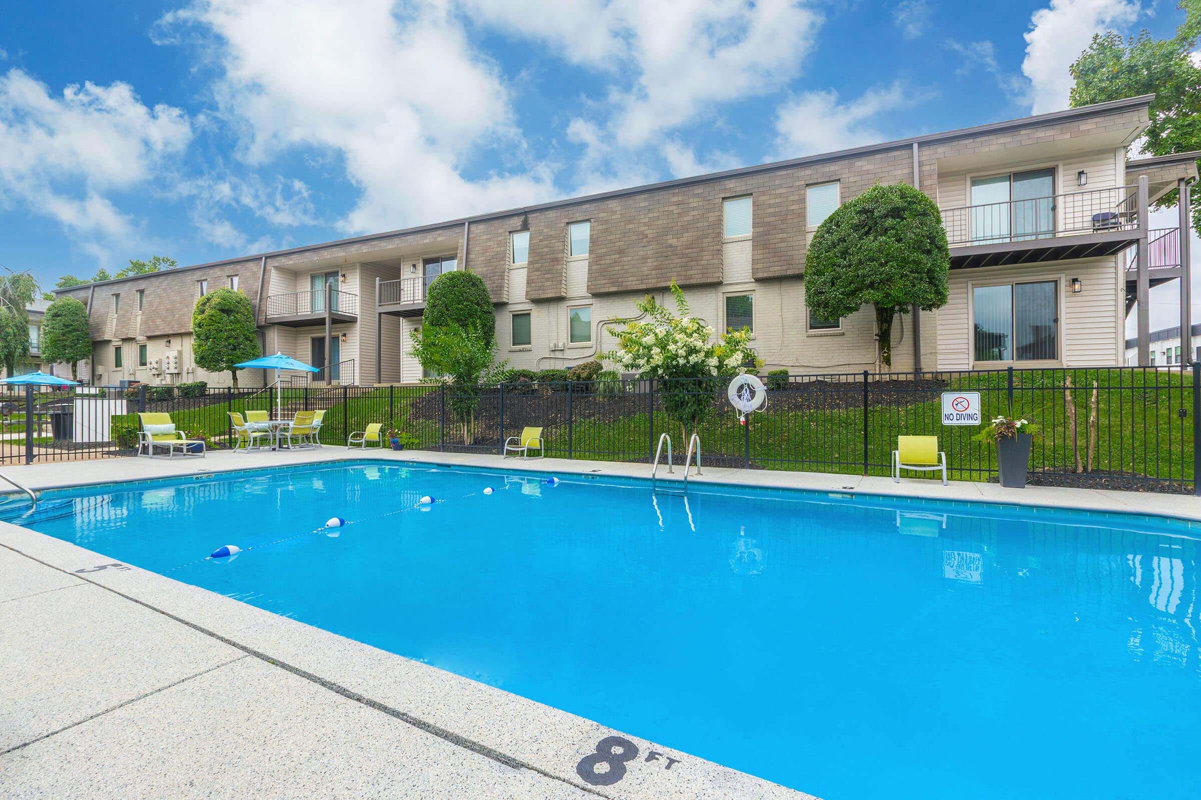 A bright outdoor pool area with clear blue water in front of a multi-story residential building. Lounge chairs and umbrellas are arranged around the pool, and there are well-maintained green shrubs and grass in the background. The pool is marked with a depth sign indicating 8 feet.