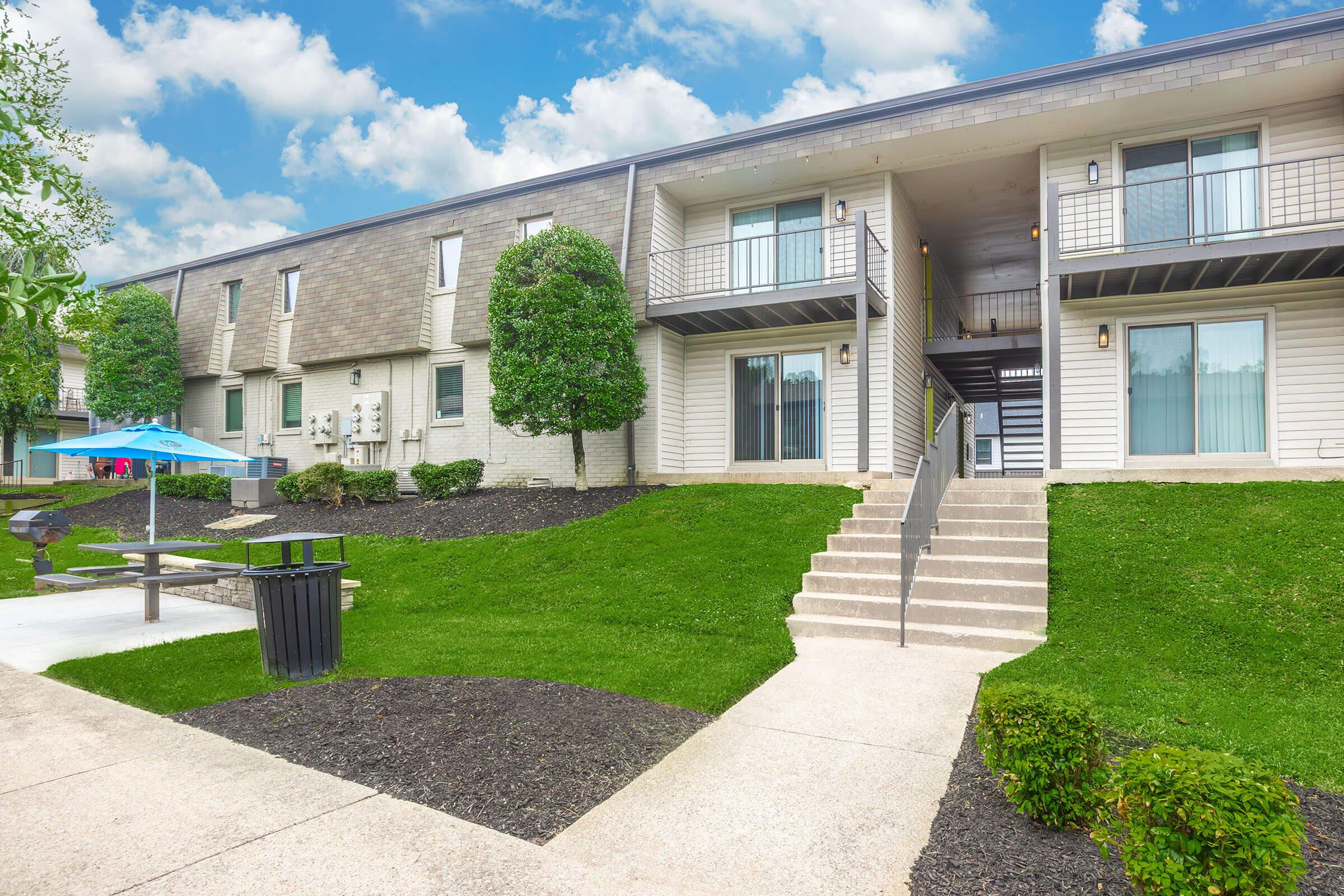 A multi-unit residential building with a light-colored exterior, featuring stairs leading to upper-level apartments. Green lawn areas and small trees are present, along with a picnic table and trash bin. The sky is partly cloudy, giving a bright, inviting atmosphere.