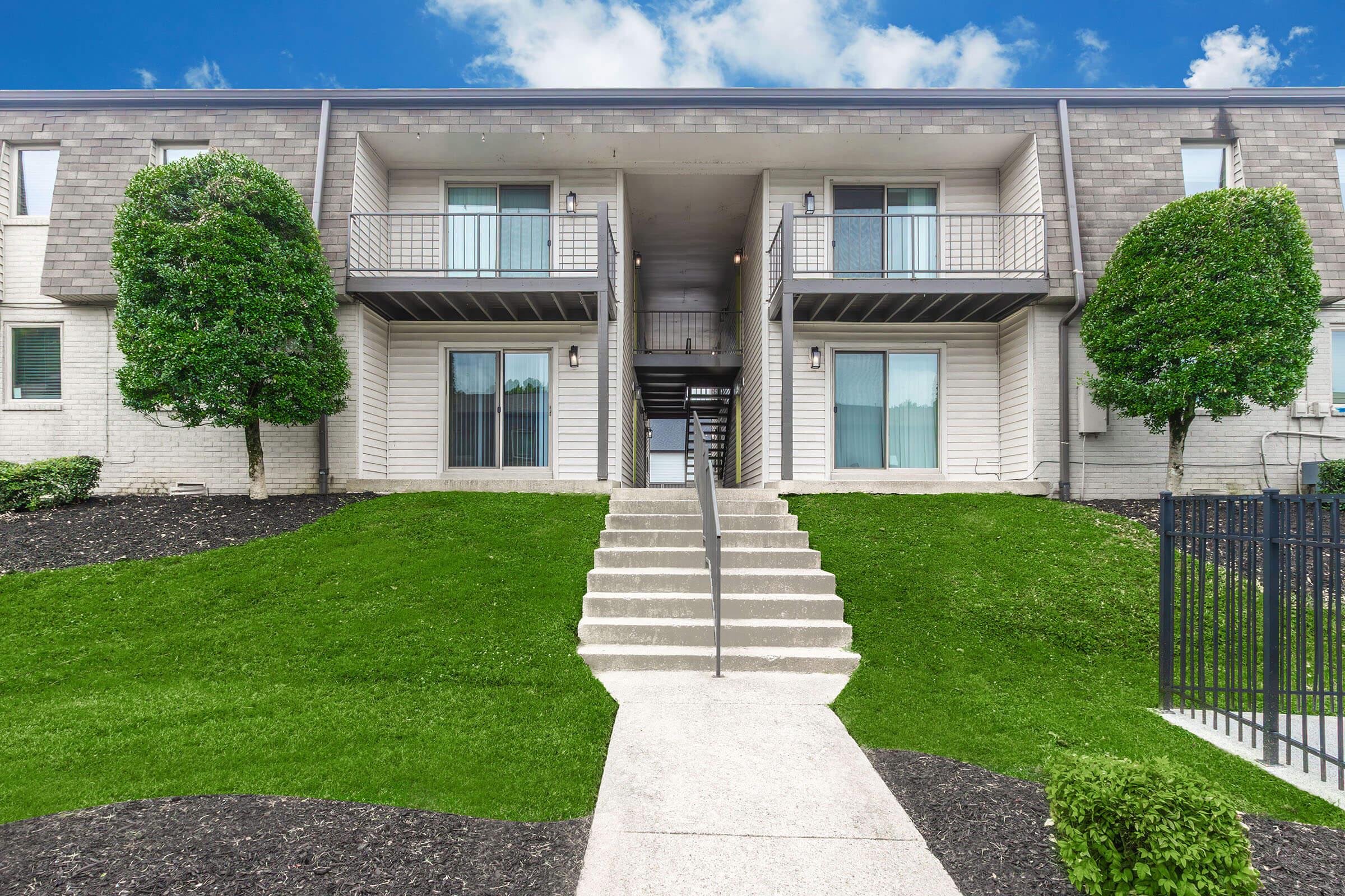 A two-story apartment building featuring a green lawn, neatly trimmed bushes, and stair steps leading to the entrance. The facade includes balconies and large windows. The sky is partly cloudy, contributing to a bright, inviting atmosphere.