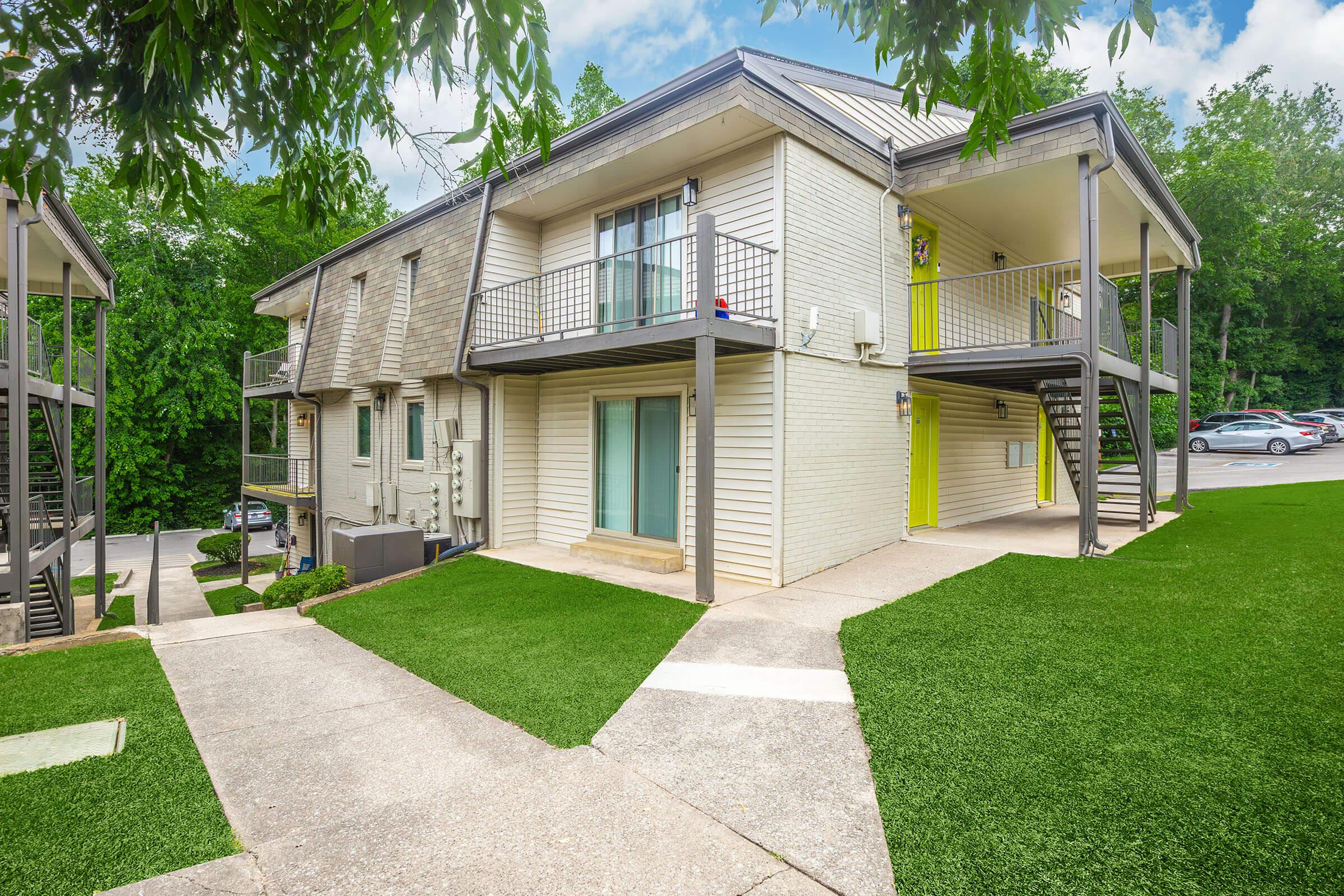 A view of a two-story apartment building with light-colored siding and green landscaping. The entrance has a staircase leading to the upper level, and there are balconies on both floors. The pathway is paved and flanked by artificial turf. Trees and a parking area are visible in the background.