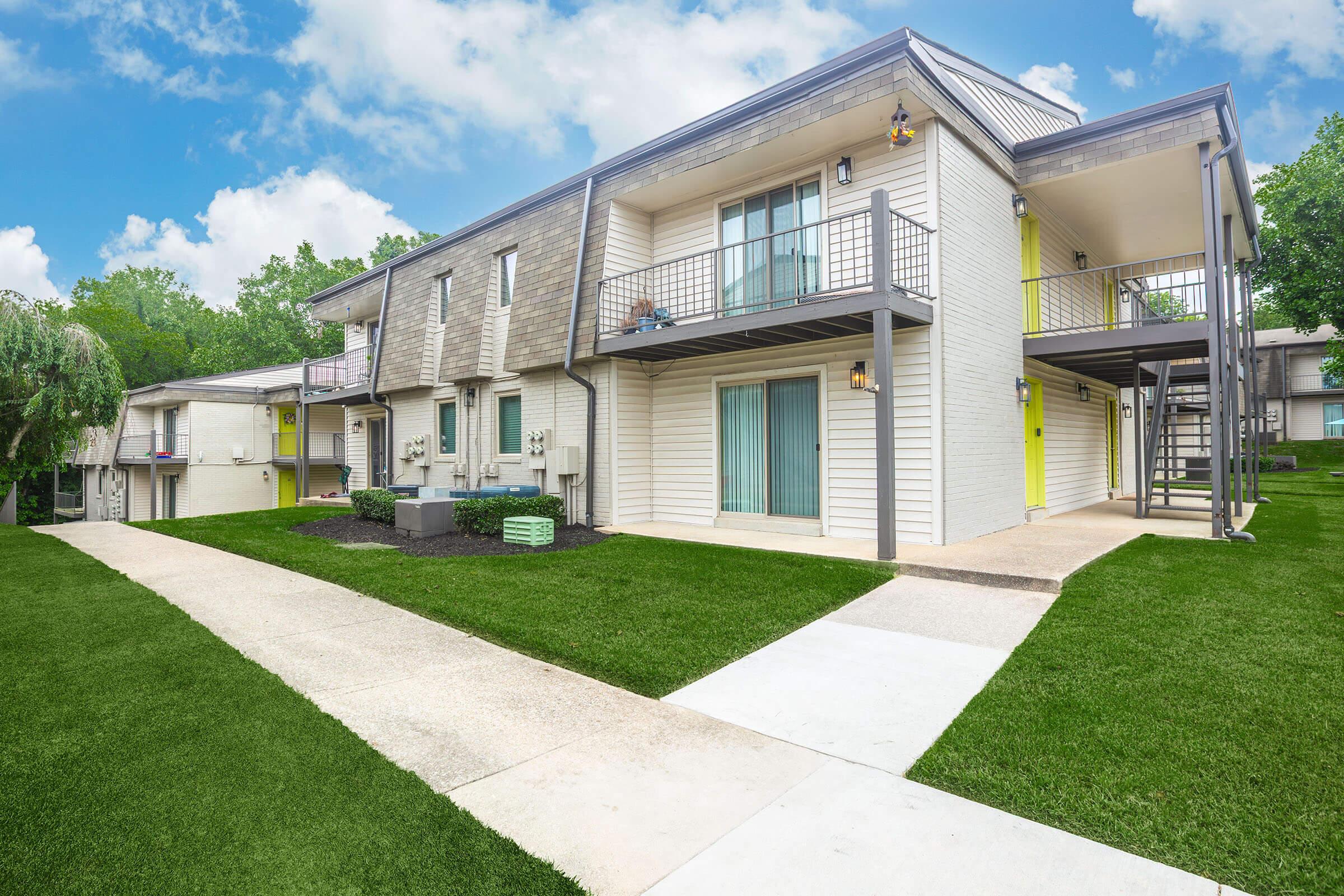 A modern residential building featuring two levels with balconies. The exterior is predominantly light-colored with some bright yellow accents. A well-maintained lawn surrounds the building, and a concrete walkway leads to the entrance, set against a backdrop of blue skies and fluffy clouds.