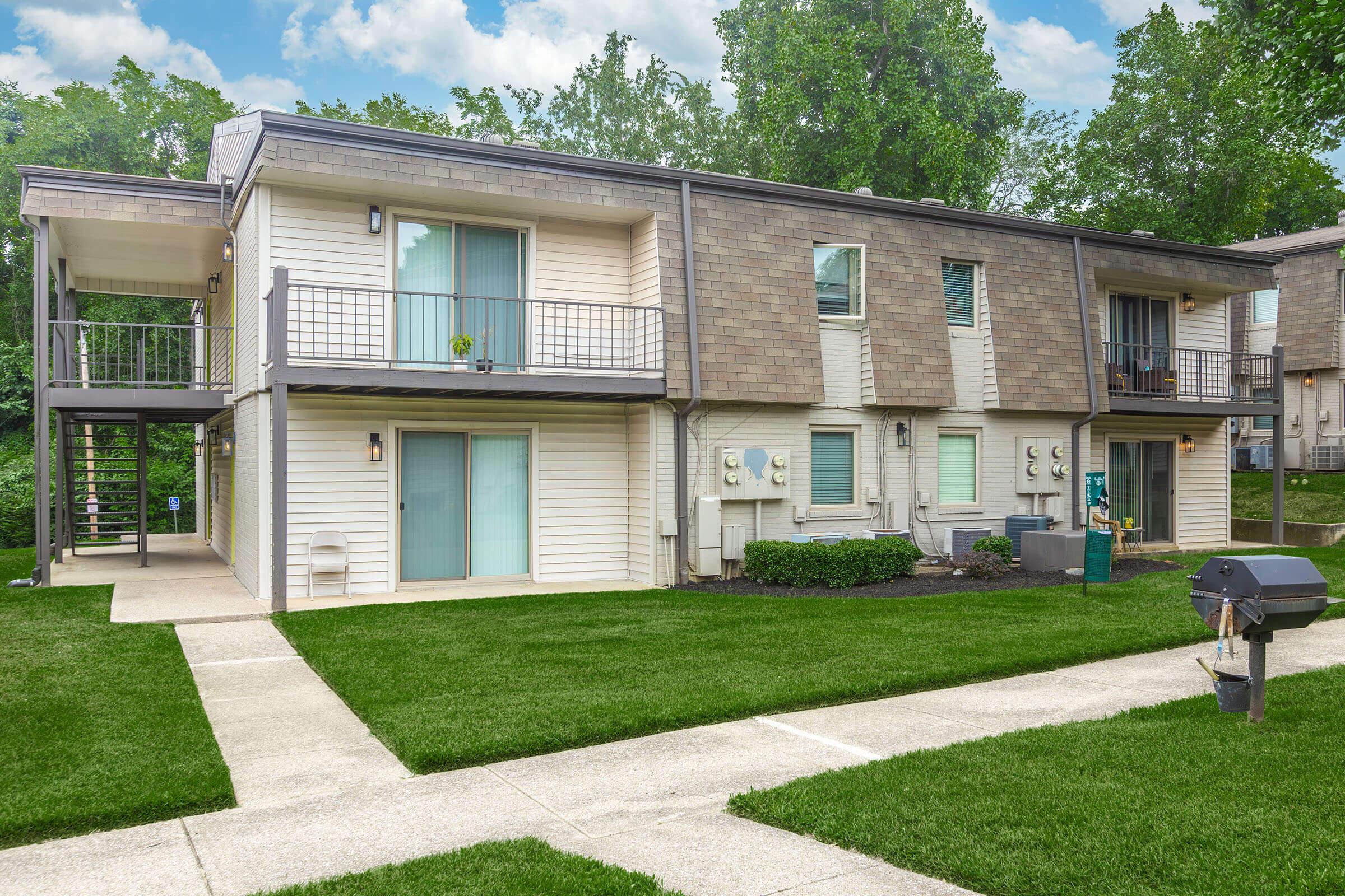 A two-story apartment building with a light-colored exterior and shingled roof. The lower level features balconies and several air conditioning units. The surrounding area is well-maintained with green grass and a concrete pathway leading to the entrance. There is a grill on the side of the building.