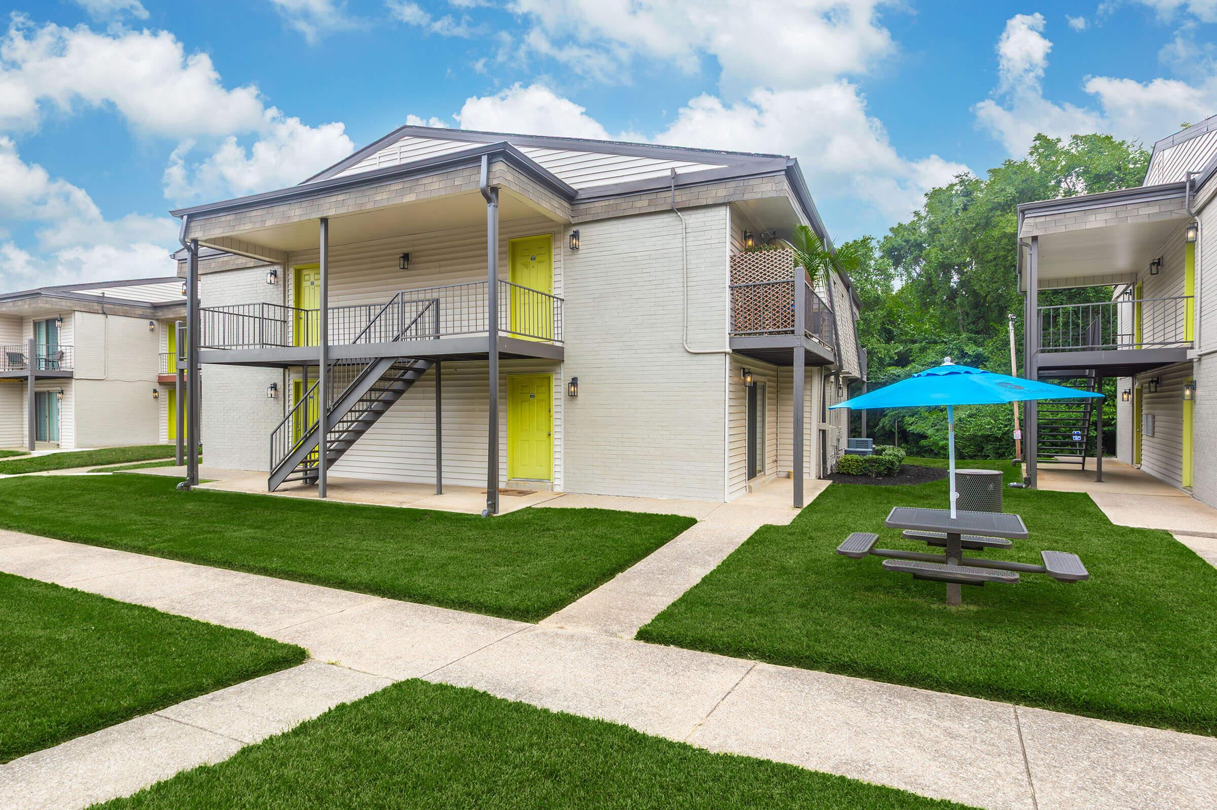 A view of a multi-unit apartment complex featuring two-story buildings with yellow accents. There are well-maintained lawns, a picnic table, and an umbrella near the walkway. The sky is clear with some clouds, creating a bright and inviting atmosphere.