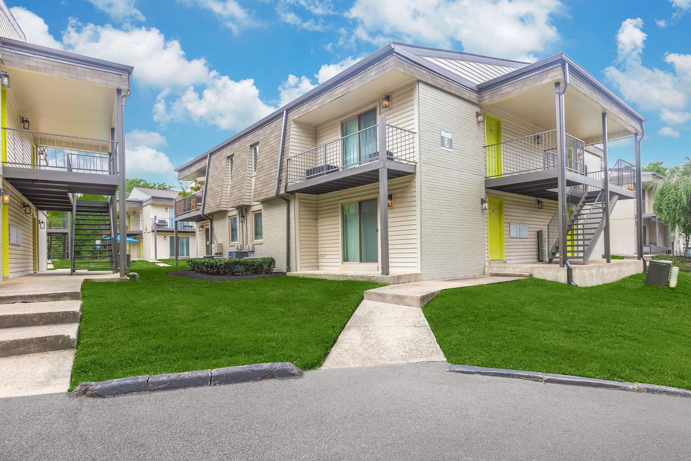 Modern two-story apartment complex with balconies, featuring light-colored brick and vibrant green accents. The landscape includes well-maintained grass and walkways leading to the entrances. Blue sky with fluffy clouds can be seen in the background, creating a pleasant atmosphere.