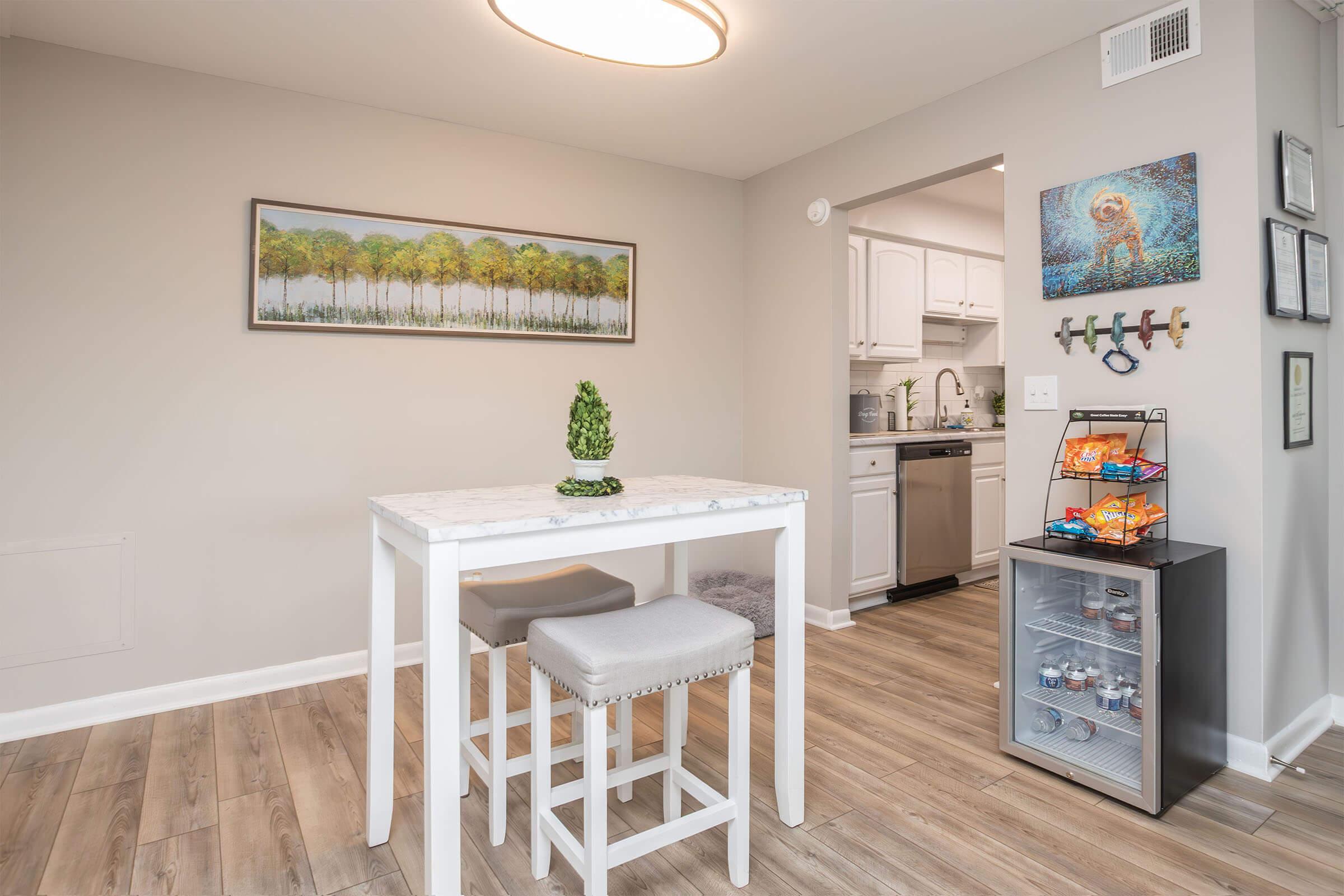 A bright kitchen area featuring a small white dining table with two gray stools. A decorative plant sits on the table. In the background, there is a kitchen with white cabinets and a light-colored wall. A colorful artwork of trees hangs on the wall, and a shelving unit displays snacks and drinks.