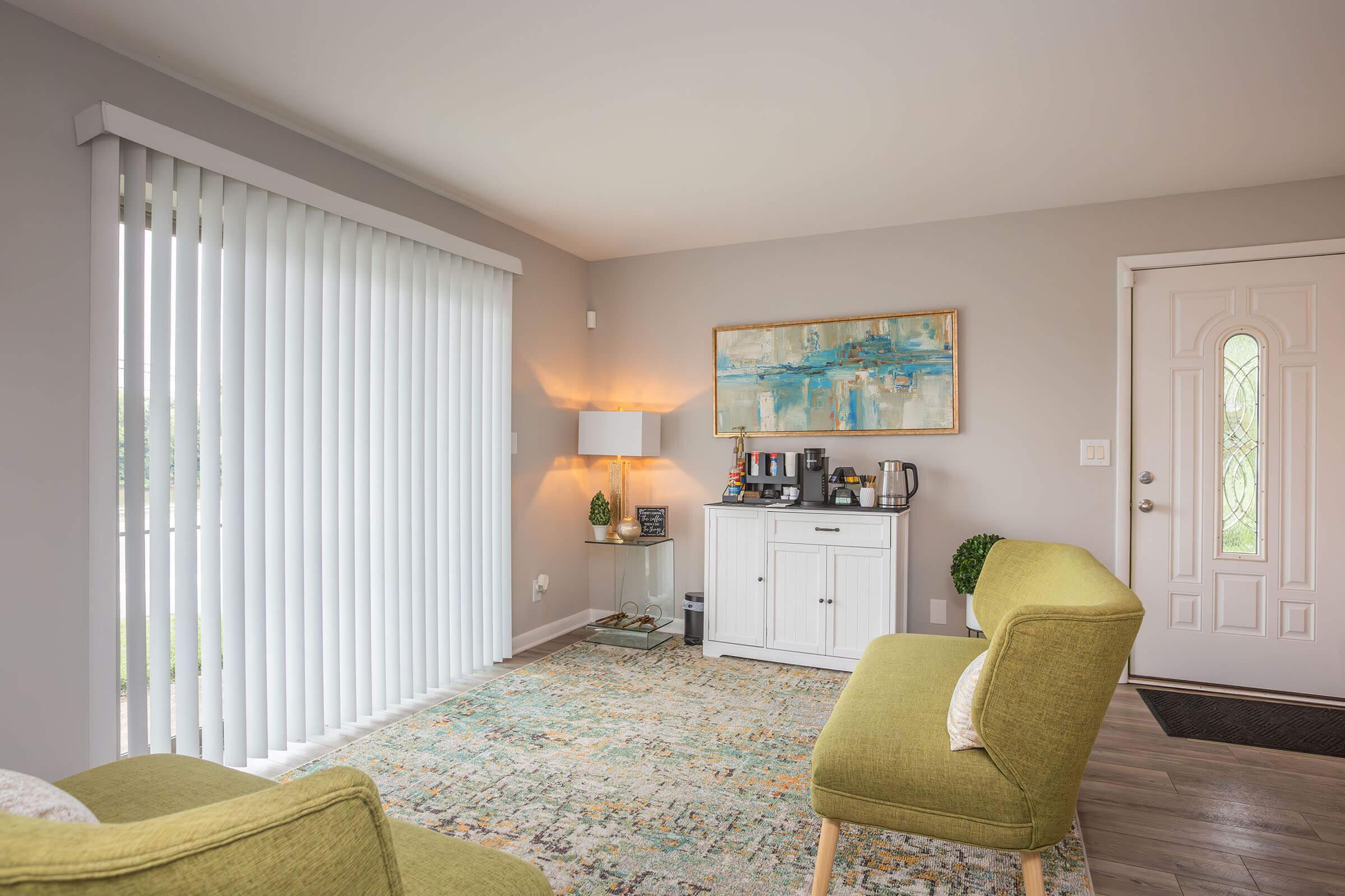 A cozy living room featuring two green chairs, a light-colored area rug, and a white cabinet against a grey wall. A lamp and decorative items are placed on the cabinet, and sheer vertical blinds cover the window. A door with a glass inset leads outside, adding natural light to the space.