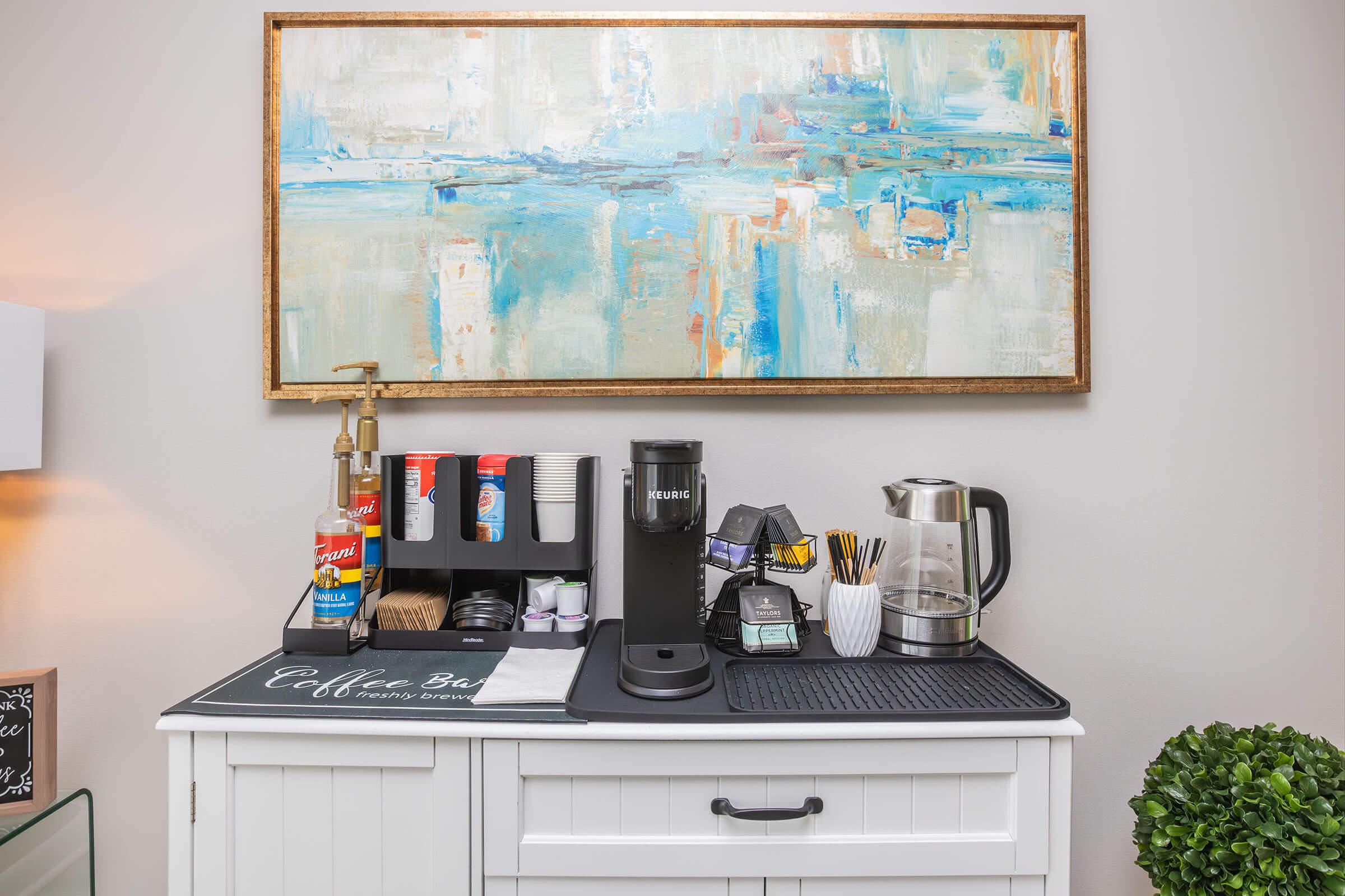 A stylish coffee bar setup featuring various coffee-making equipment. There’s a coffee machine, kettle, and cups on a white cabinet, complemented by a colorful abstract painting on the wall above. The area is organized with decor items, creating a cozy atmosphere for enjoying coffee.