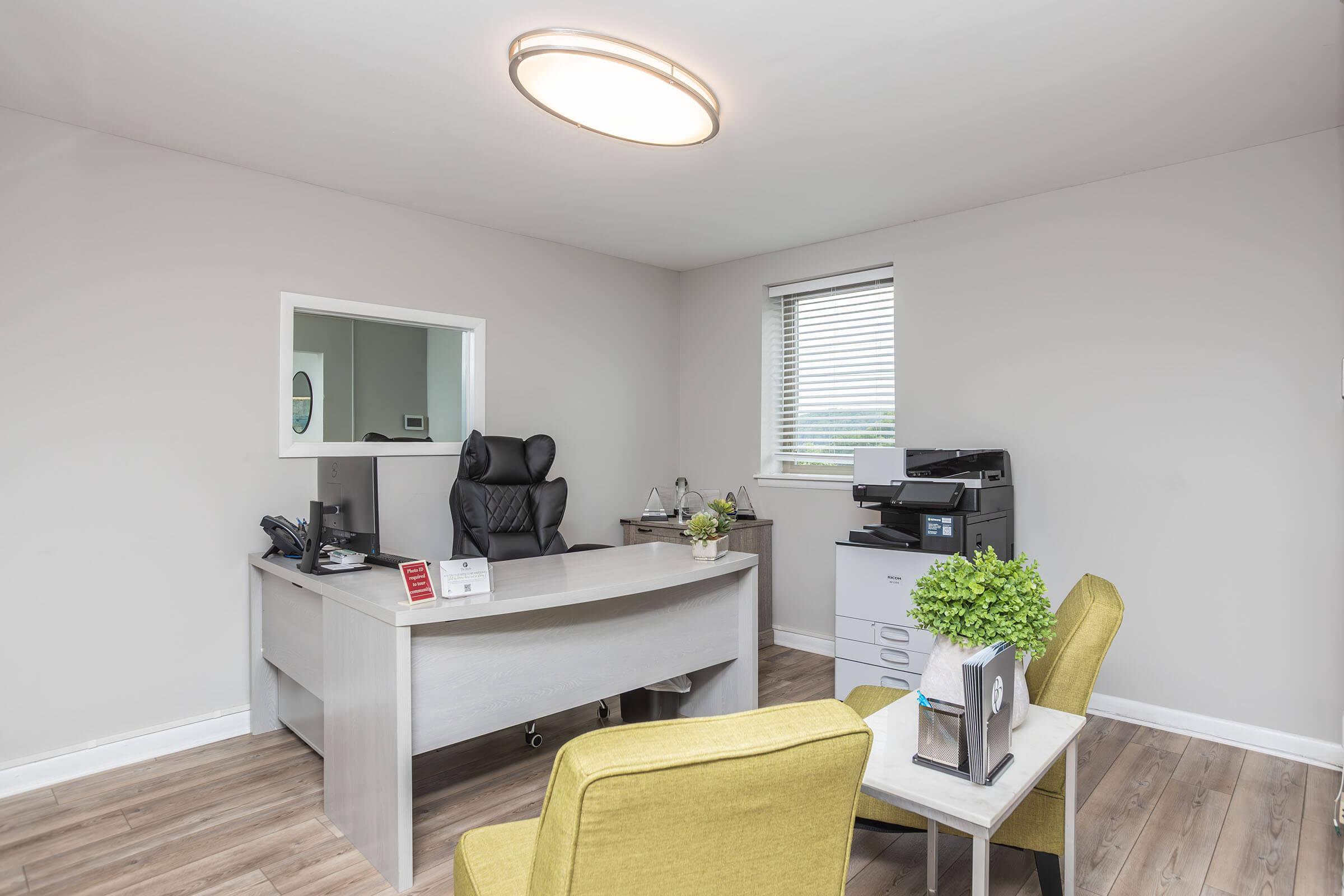 A modern office space featuring a sleek desk with a computer monitor, an ergonomic black chair, and a printer. Two bright yellow chairs are positioned nearby, with a small plant on the desk and another on a shelf. Natural light streams in through a window, enhancing the neutral-colored walls and wooden floor.