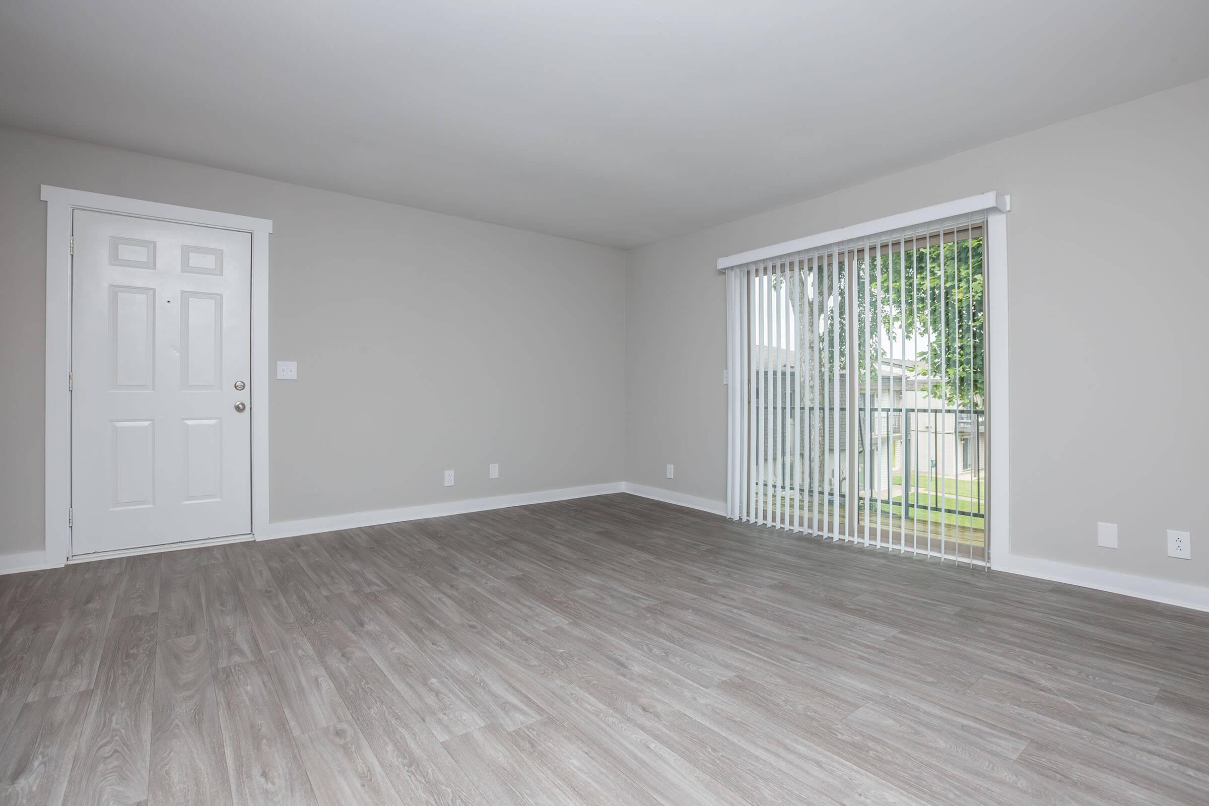 A vacant living room with light gray walls and wide, light-colored vinyl flooring. There is a white door on the left and a sliding glass door on the right that leads outside, covered with vertical blinds. The space is well-lit and has a clean, modern appearance.