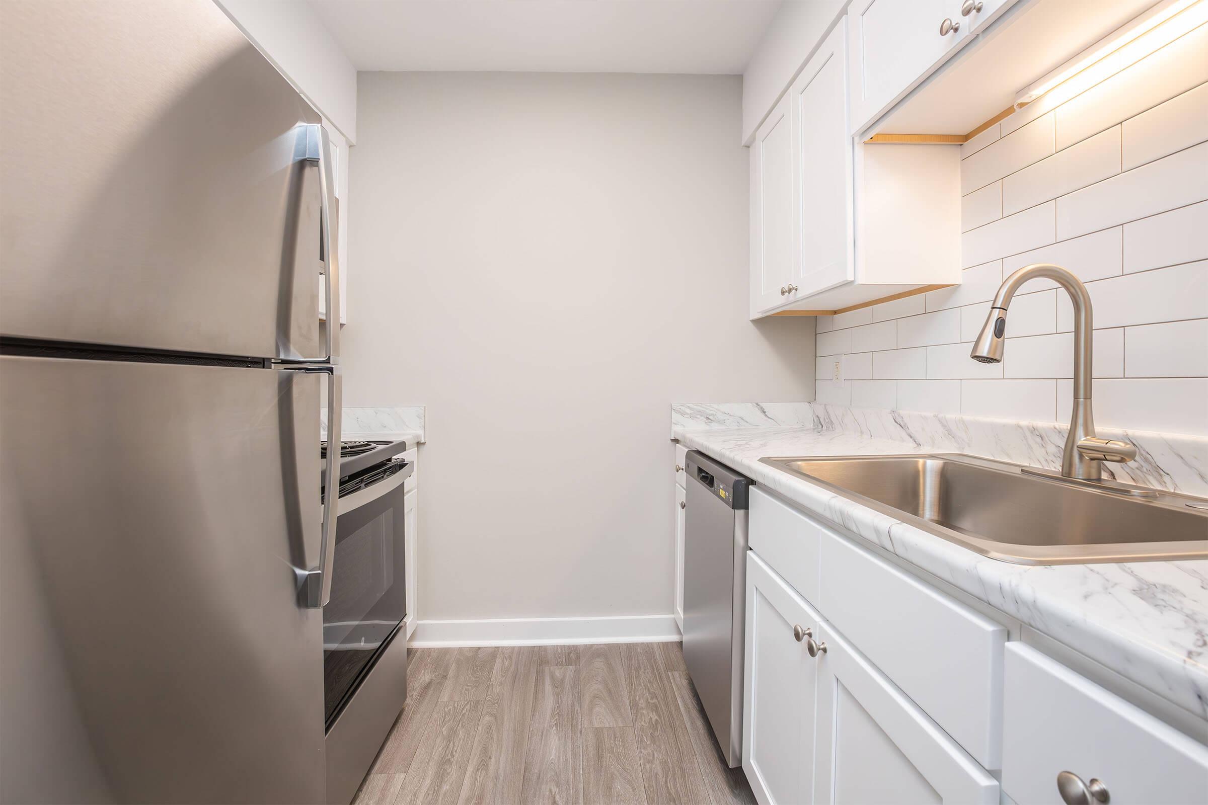 Modern kitchen featuring stainless steel appliances, including a refrigerator, oven, and dishwasher. The white cabinets complement the marble countertop, while the backsplash consists of white subway tiles. The space has a bright, clean aesthetic with grey wood-like flooring.