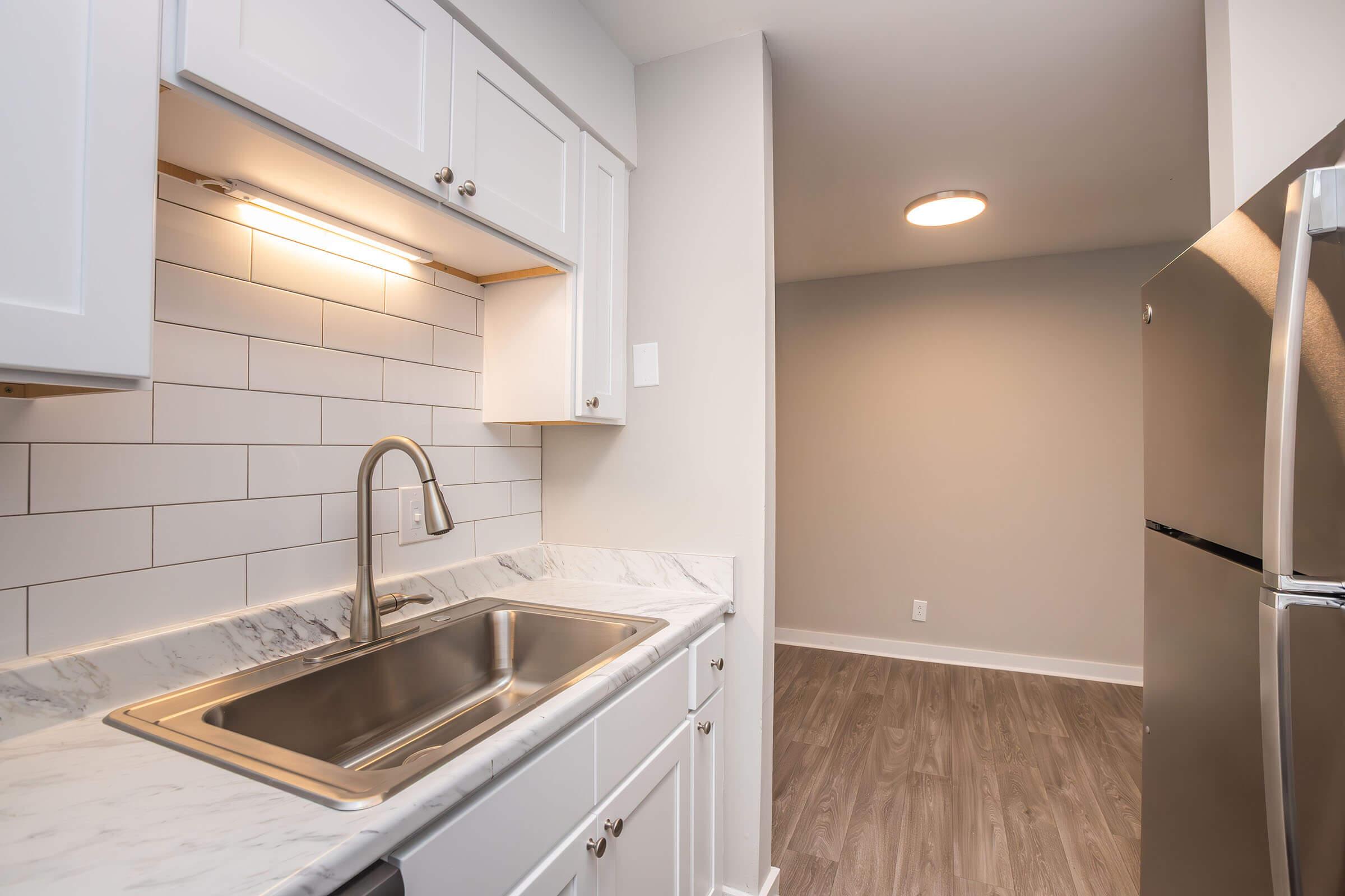 A modern kitchen featuring white cabinetry, a stainless steel sink with a single faucet, and a marble countertop. The backsplash consists of white tiles. To the right, there's a stainless steel refrigerator and a light-colored wall, leading to an area with gray flooring and a ceiling light fixture.