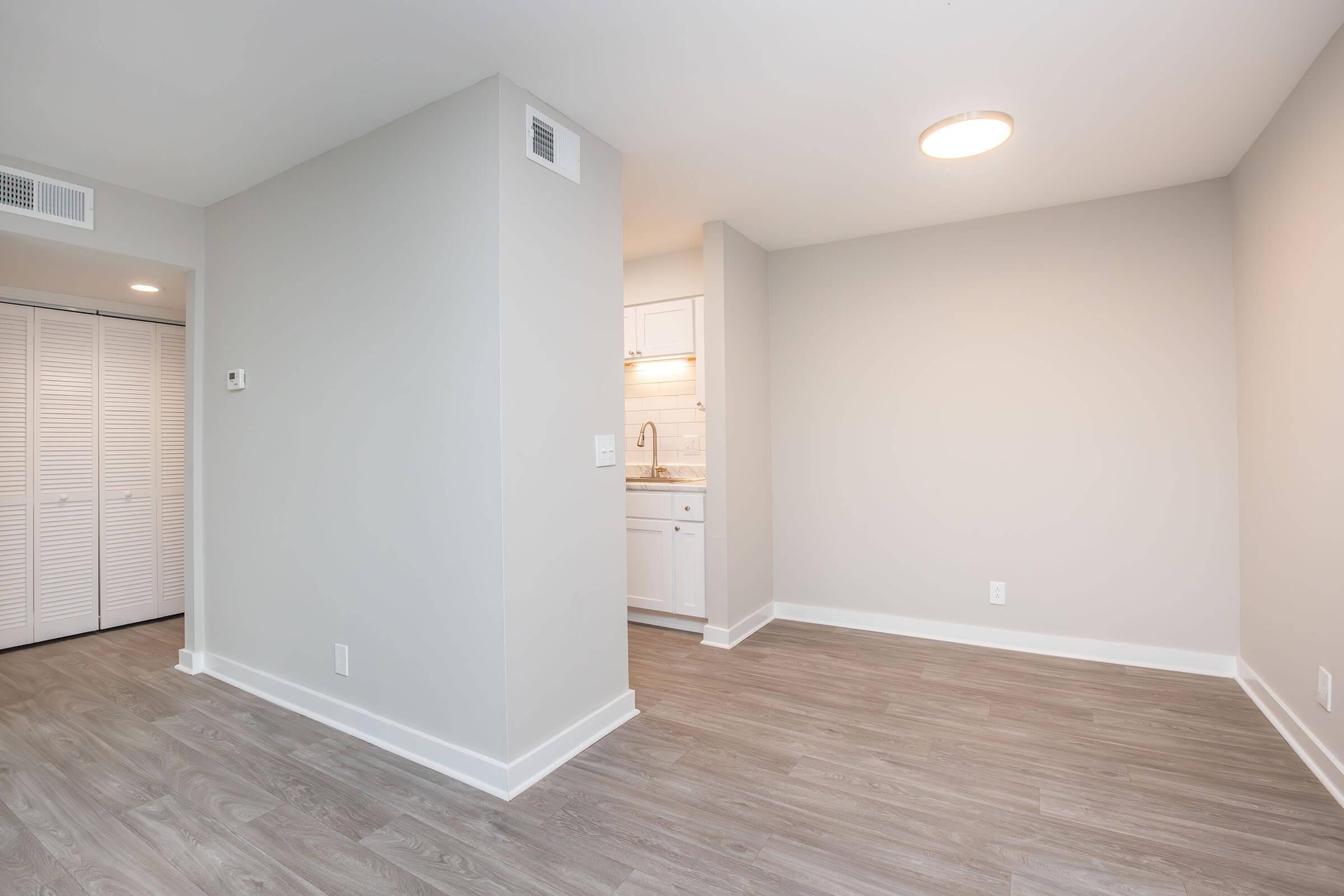A bright, unfurnished room with light gray walls and laminate flooring. The space includes a corner that opens to a kitchen area, visible with white cabinetry. There is a closet with bi-fold doors on one side, and a ceiling light fixture illuminates the room.