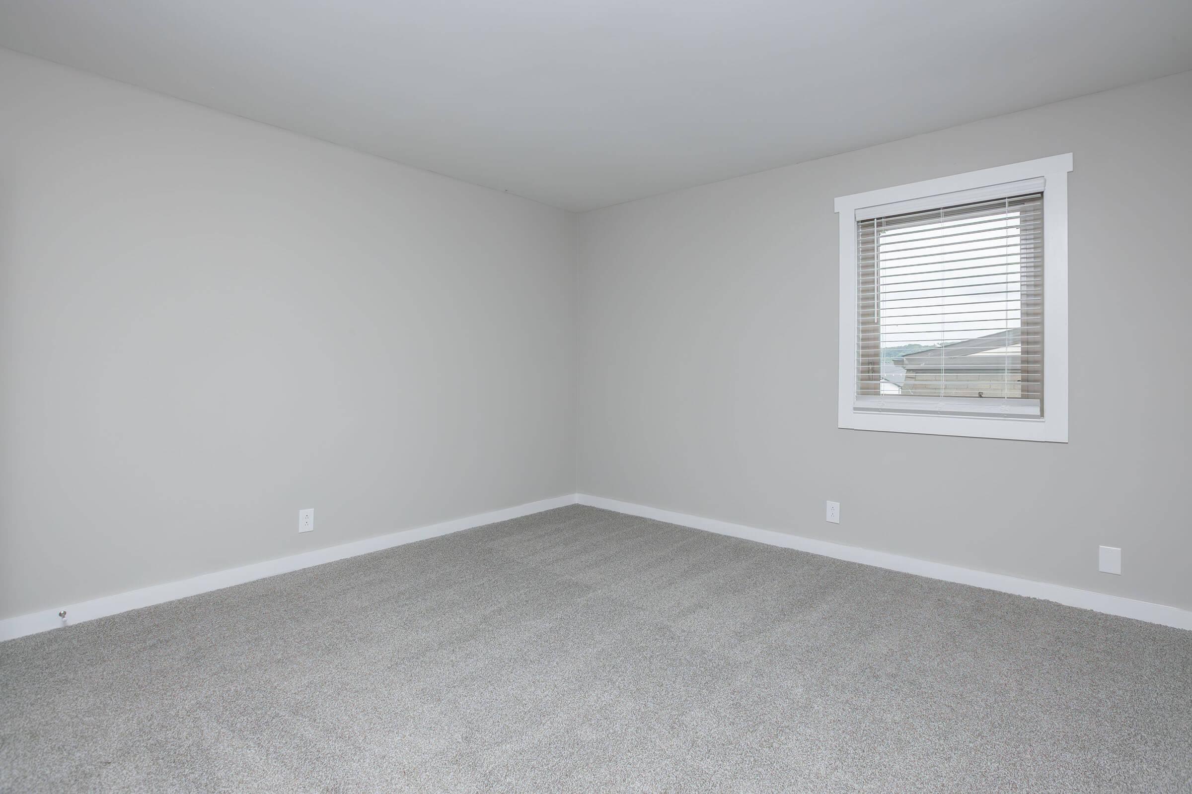 A vacant room with light gray walls and a window adorned with blinds. The carpeted floor is also light gray, creating a neutral and airy atmosphere. The space is well-lit and uncluttered, ideal for various purposes, such as a bedroom or office.