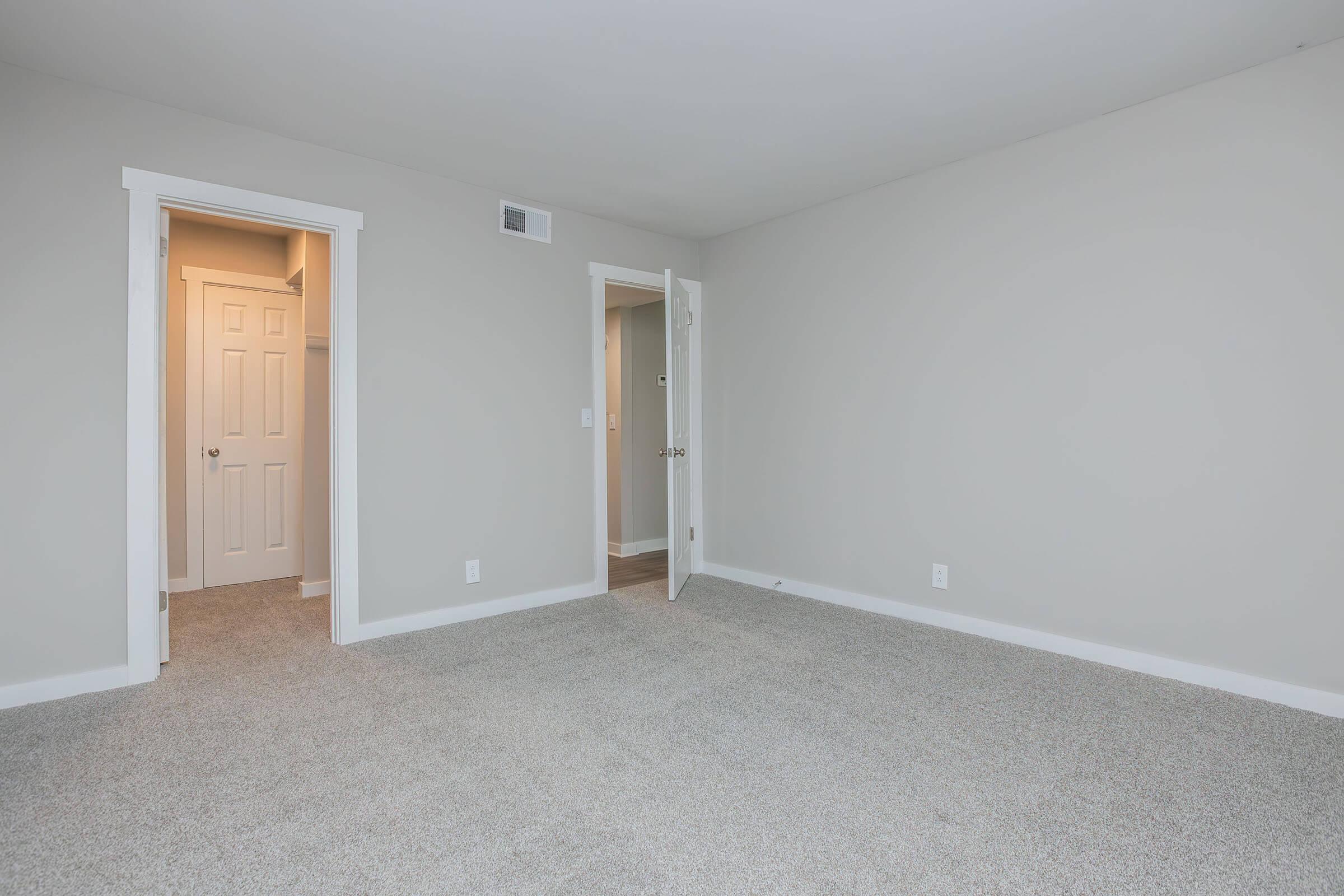 A spacious, empty room with light gray walls and plush carpet. There are two doors: one leading to a closet and another to an adjacent area. The room is well-lit, creating a bright atmosphere, and features white trim around the doors and baseboards.