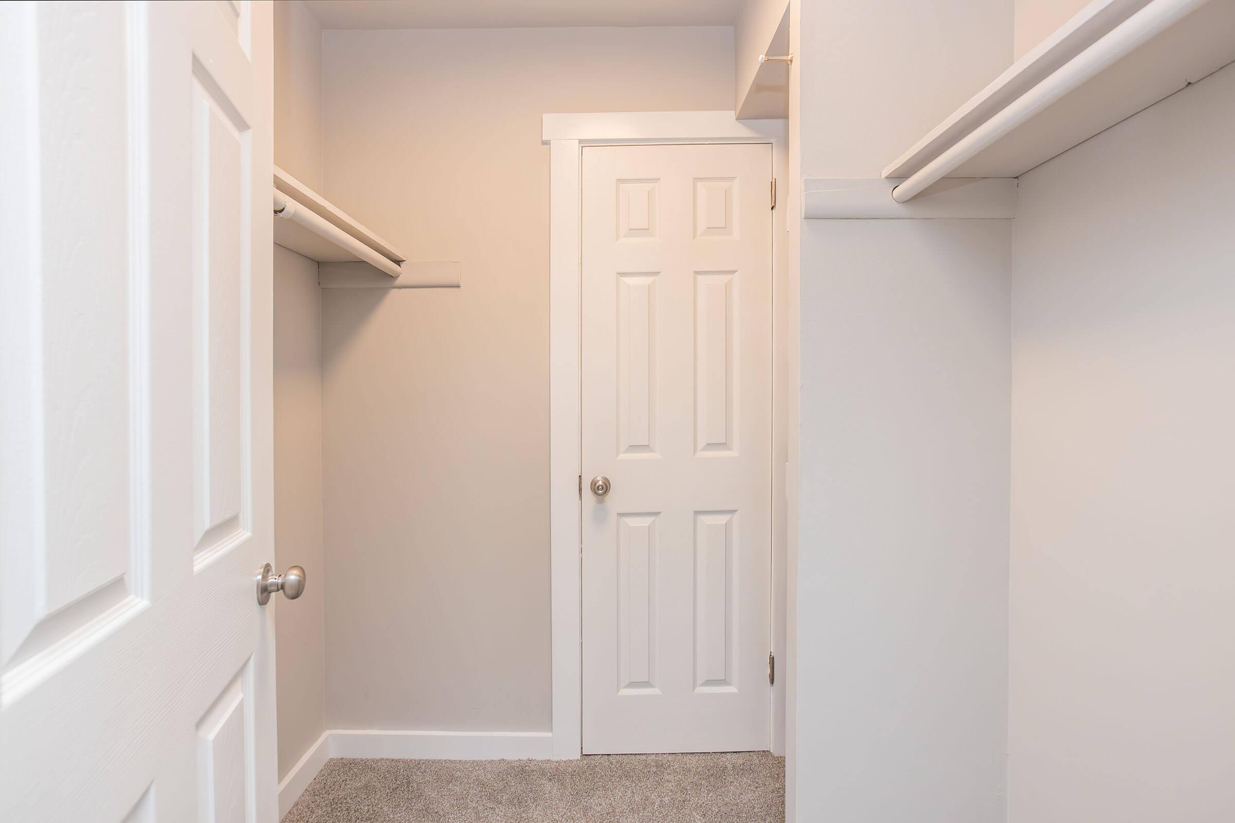 A light-colored closet space featuring white walls and shelving on either side. A closed white door is visible at the back, and the floor is carpeted in a neutral tone. The overall appearance is clean and minimalistic.