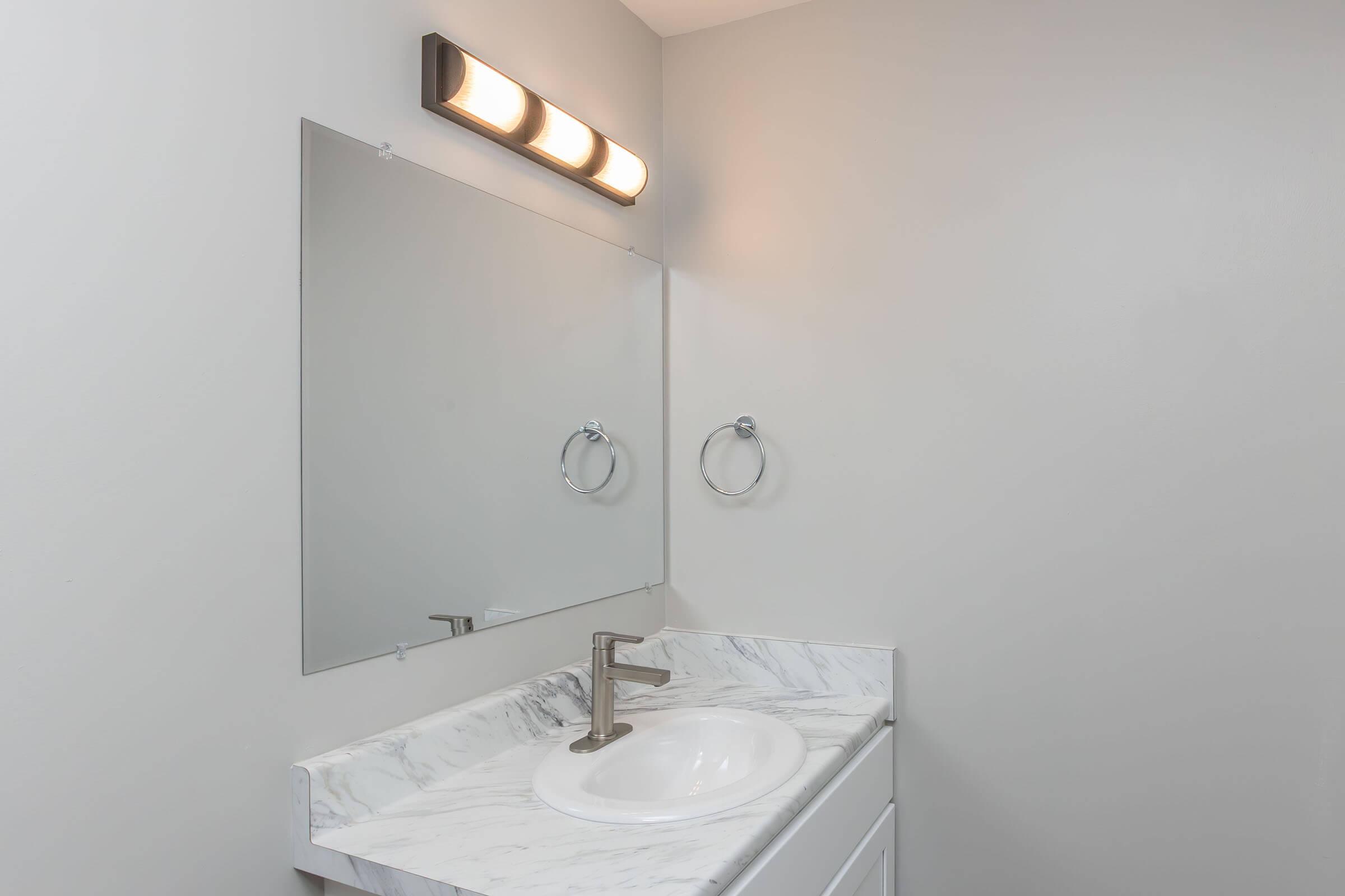 A modern bathroom vanity featuring a white marble countertop, a round sink, and a sleek faucet. Above the sink, there's a large rectangular mirror and a light fixture with four bulbs. The walls are painted gray, and there are chrome towel rings on either side of the mirror. The overall design is minimalist and contemporary.