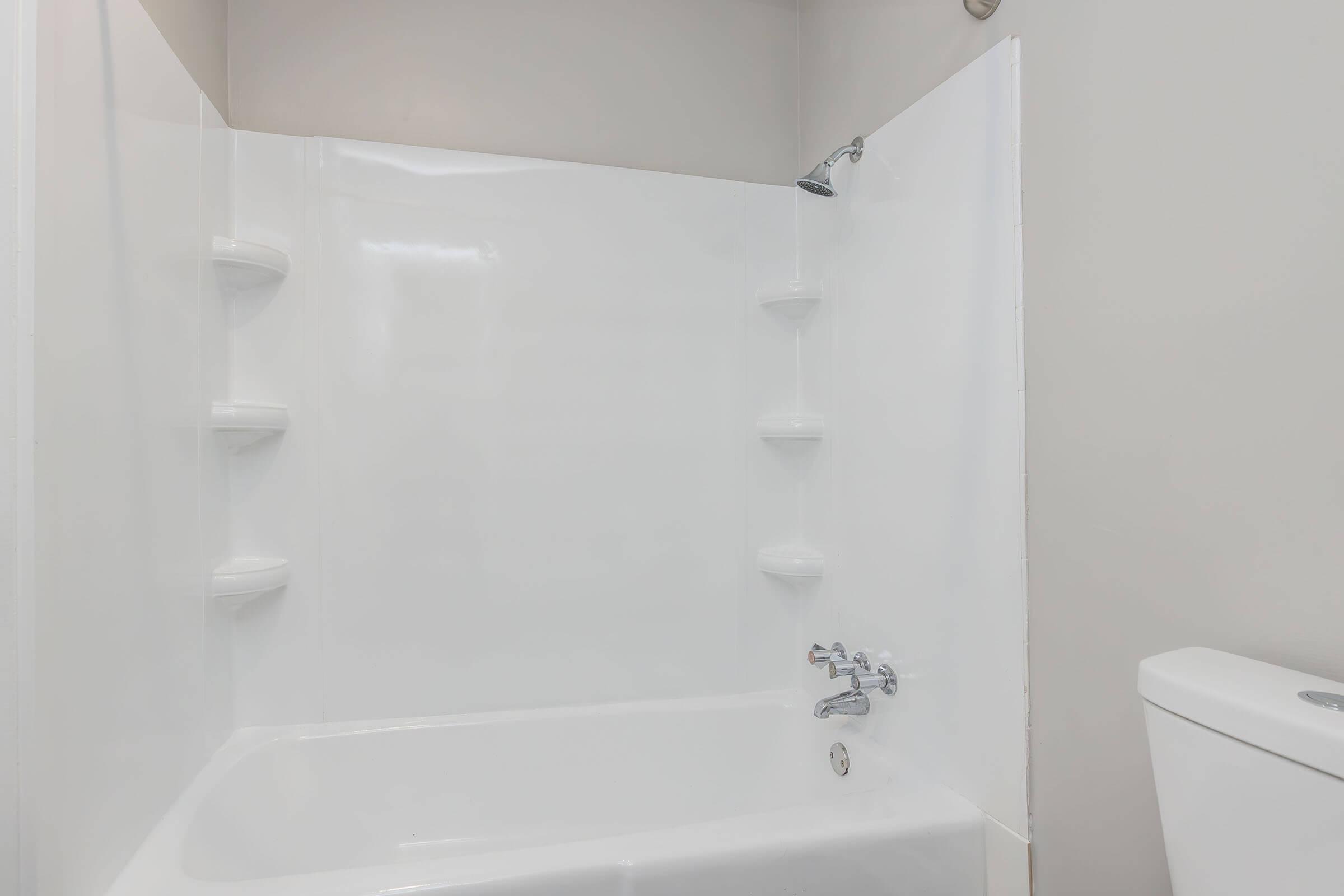 A clean, modern bathroom with a white bathtub and shower combination. The walls are smooth and white, featuring built-in shelves for toiletries. A chrome faucet and showerhead are visible, alongside a white toilet to the right. The overall aesthetic is bright and minimalistic.