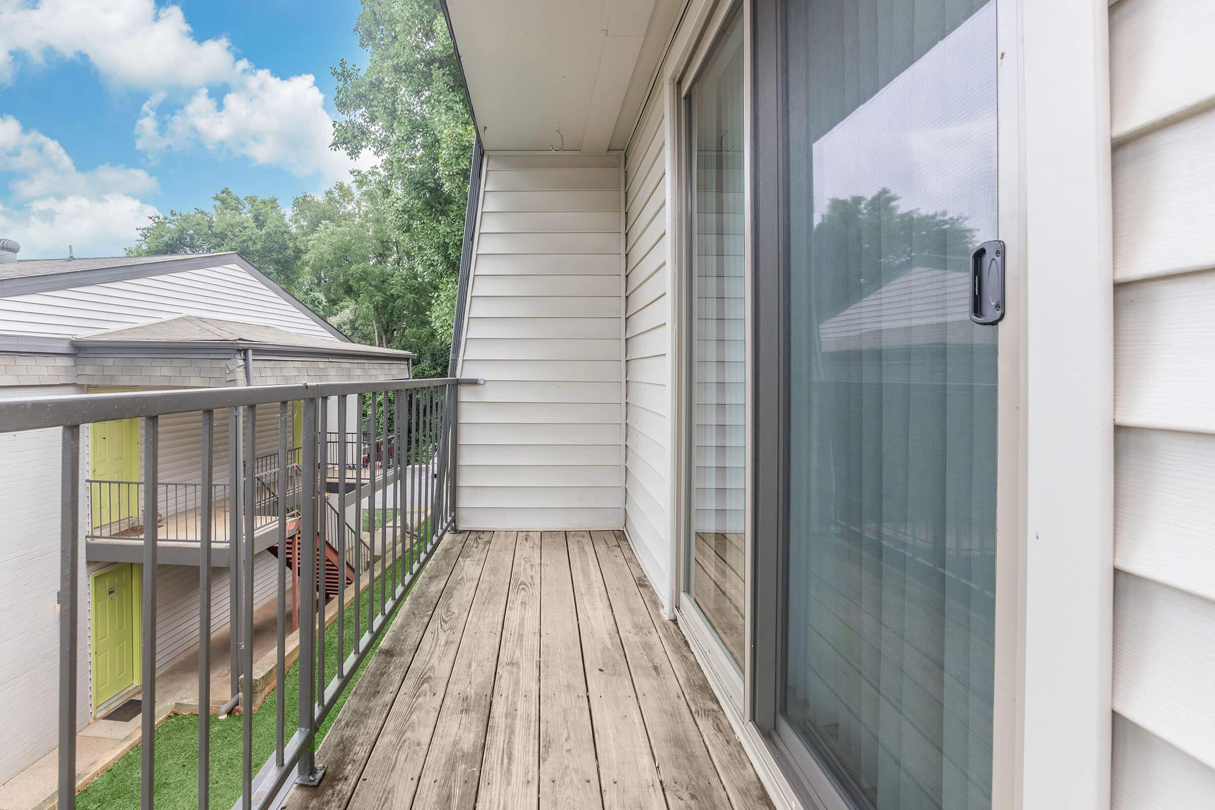 A wooden balcony with a metal railing, featuring sliding glass doors leading into a room. Surrounding the balcony are light-colored apartment buildings and green trees under a partly cloudy sky. The floor is wooden planks, and there is a grassy area visible below.