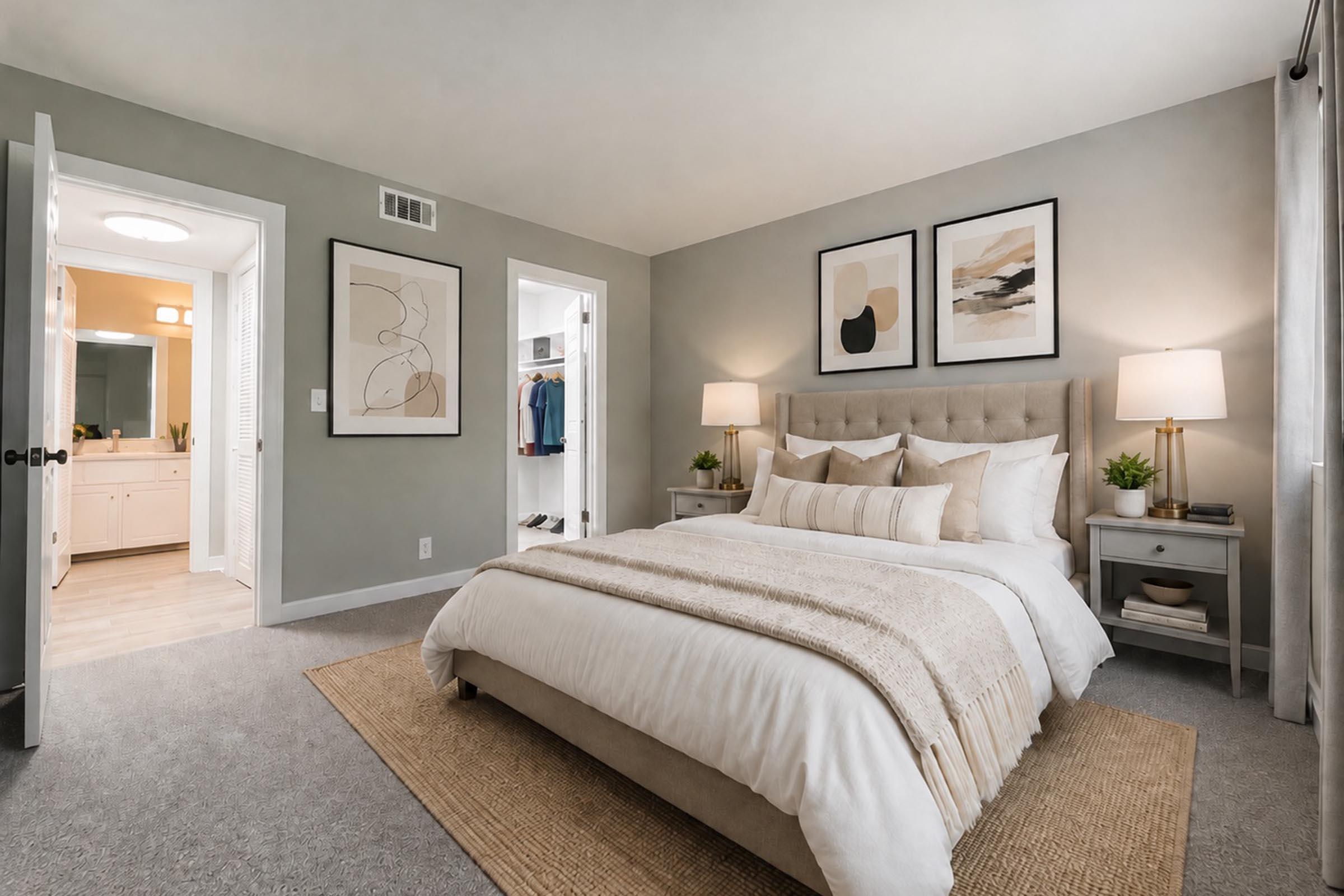 A cozy, modern bedroom featuring a large bed with multiple pillows and a textured throw blanket. Two bedside lamps illuminate the room, and artwork hangs on the walls. A doorway leads to a bathroom area, while another closet space is visible to the left. Light gray walls and a woven rug enhance the inviting atmosphere.