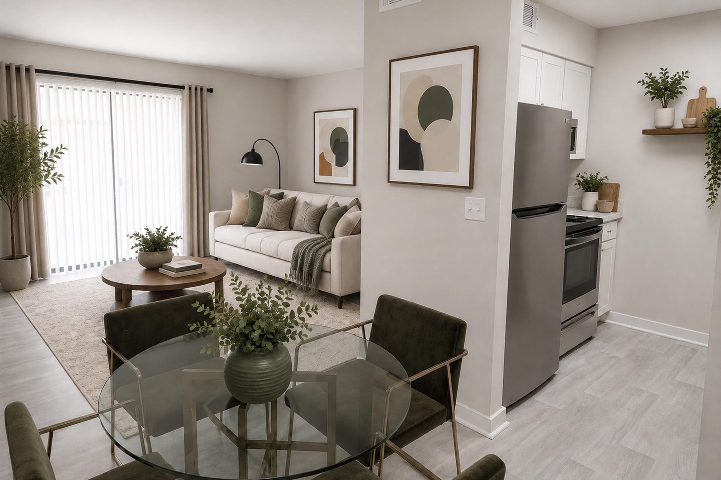 A modern living space featuring a cozy sofa with decorative pillows, a round coffee table, and a dining area with a glass table and green plants. The kitchen has stainless steel appliances, and large sliding doors lead to an outdoor space, complemented by neutral-colored walls and artwork.